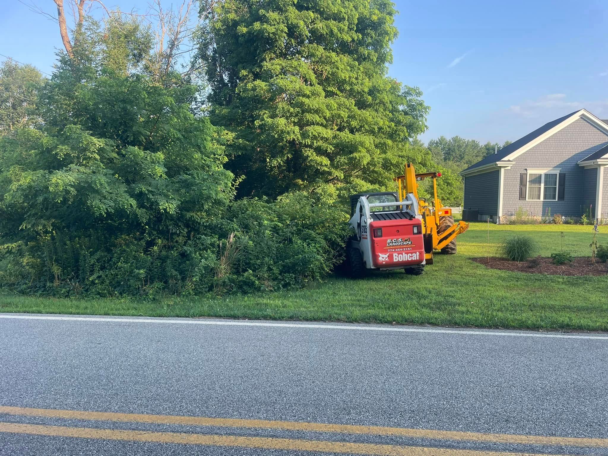 Small construction vehicle parked on grass next to a road, near a house and trees.
