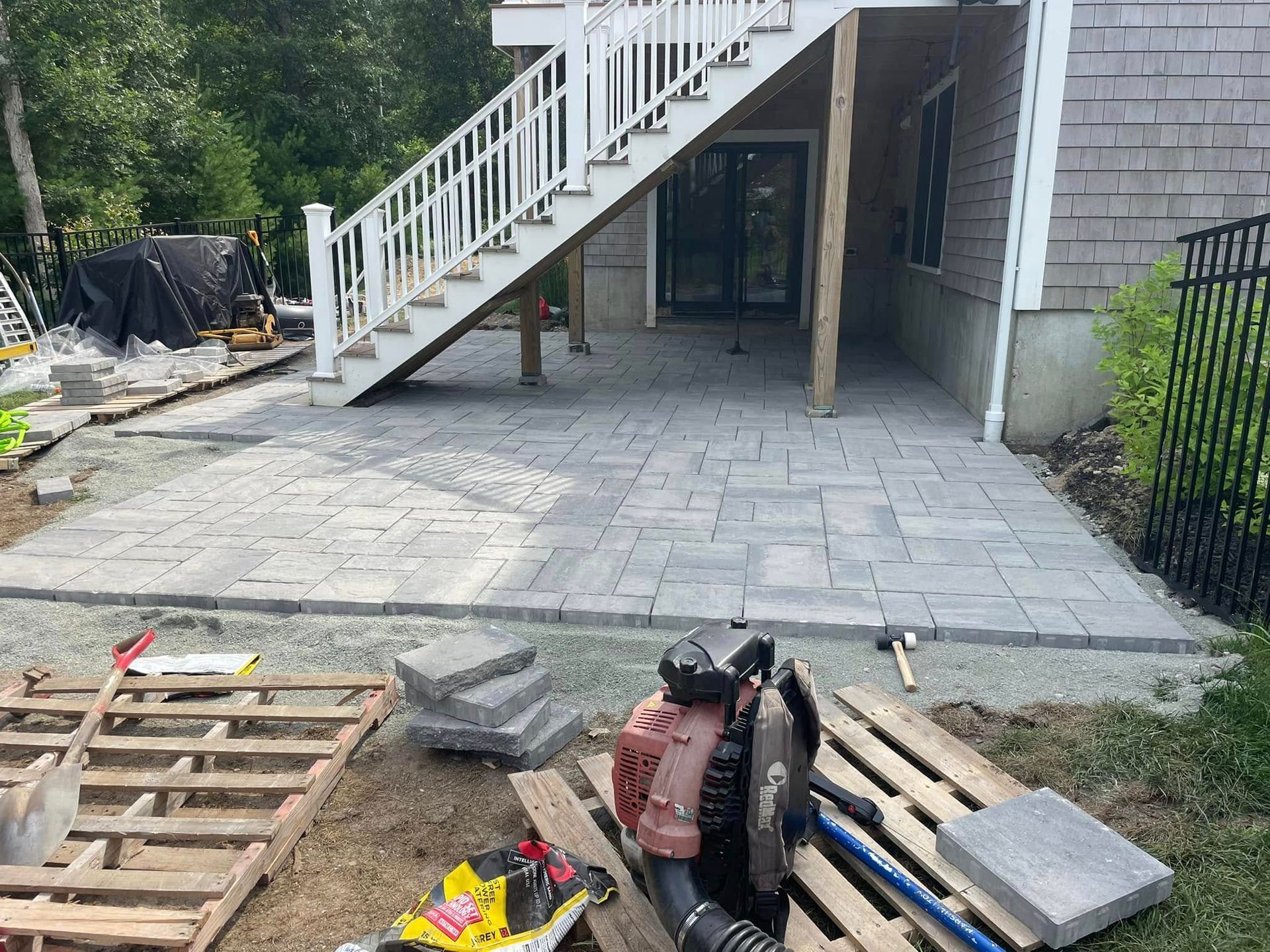 Patio construction: Gray pavers being laid under a white staircase. Tools and materials surround.