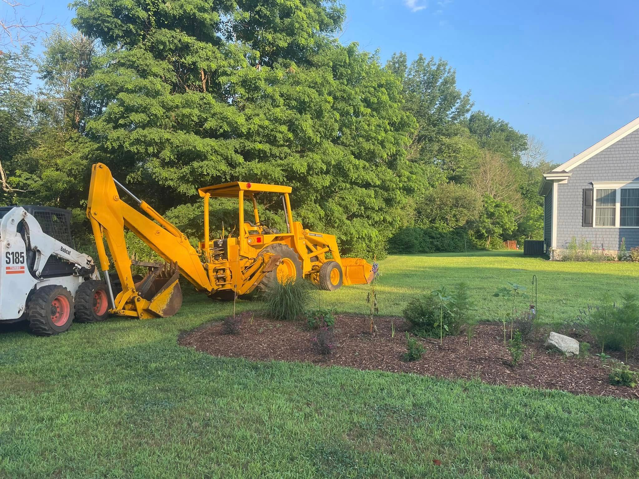 Yellow backhoe and white Bobcat on a lawn with mulch beds near a house and trees.