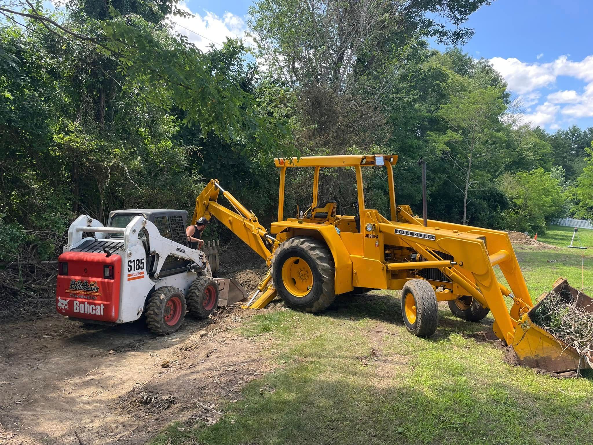 Yellow backhoe and white Bobcat skid steer digging dirt in a grassy area.