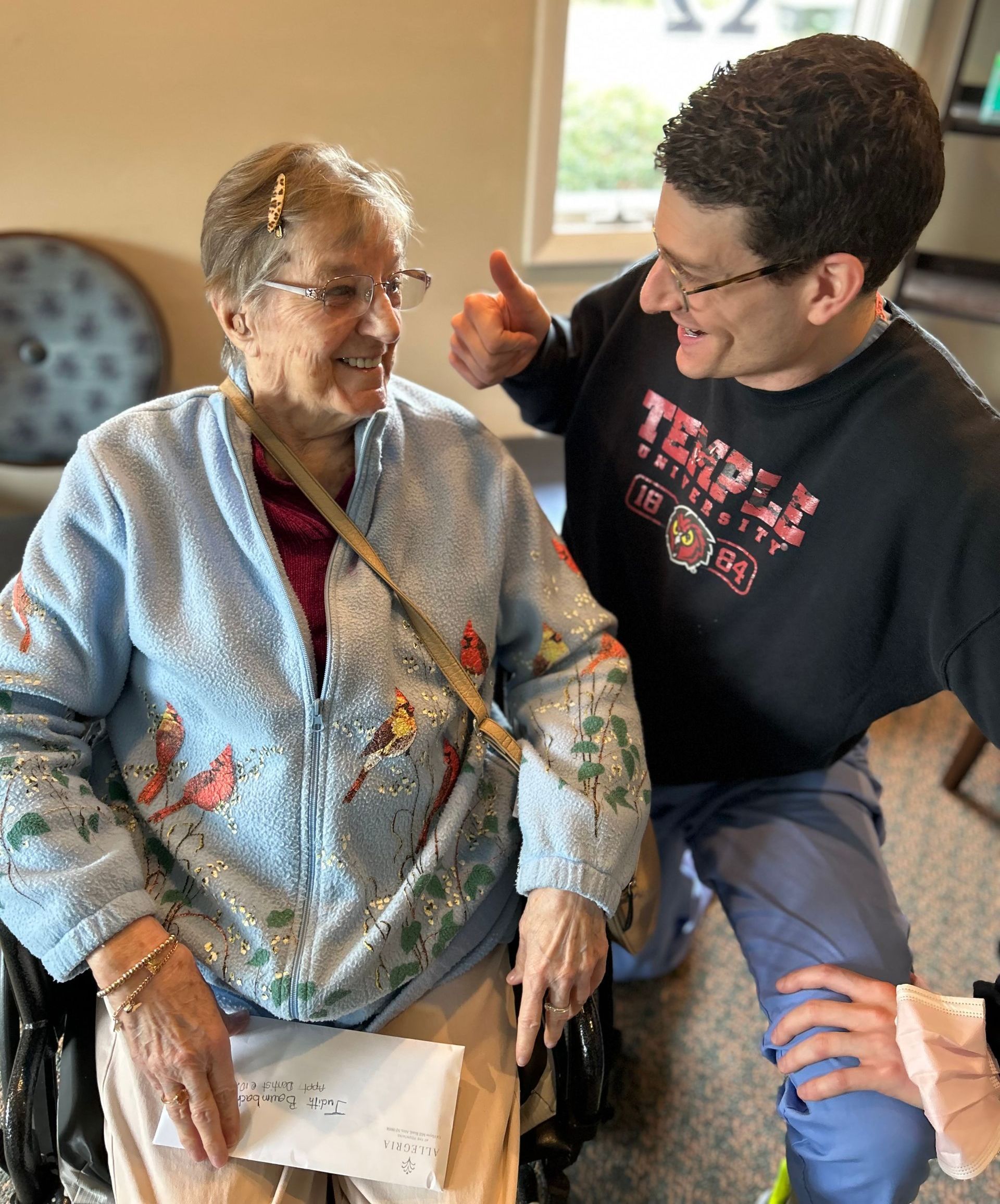 A man is kneeling down next to an elderly woman in a wheelchair and giving her a thumbs up.