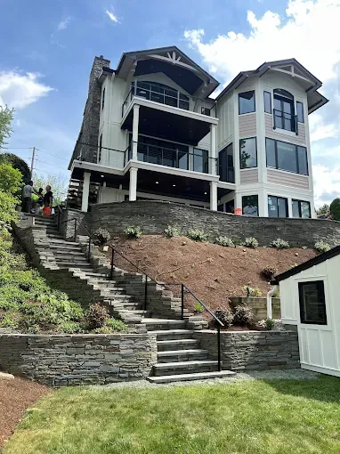 Large modern house on a steep hillside with stone steps, retaining wall, and blue sky above.