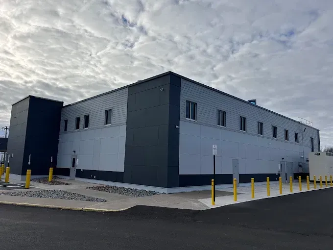 Gray industrial building with dark trim, yellow bollards, and a paved lot under a cloudy sky