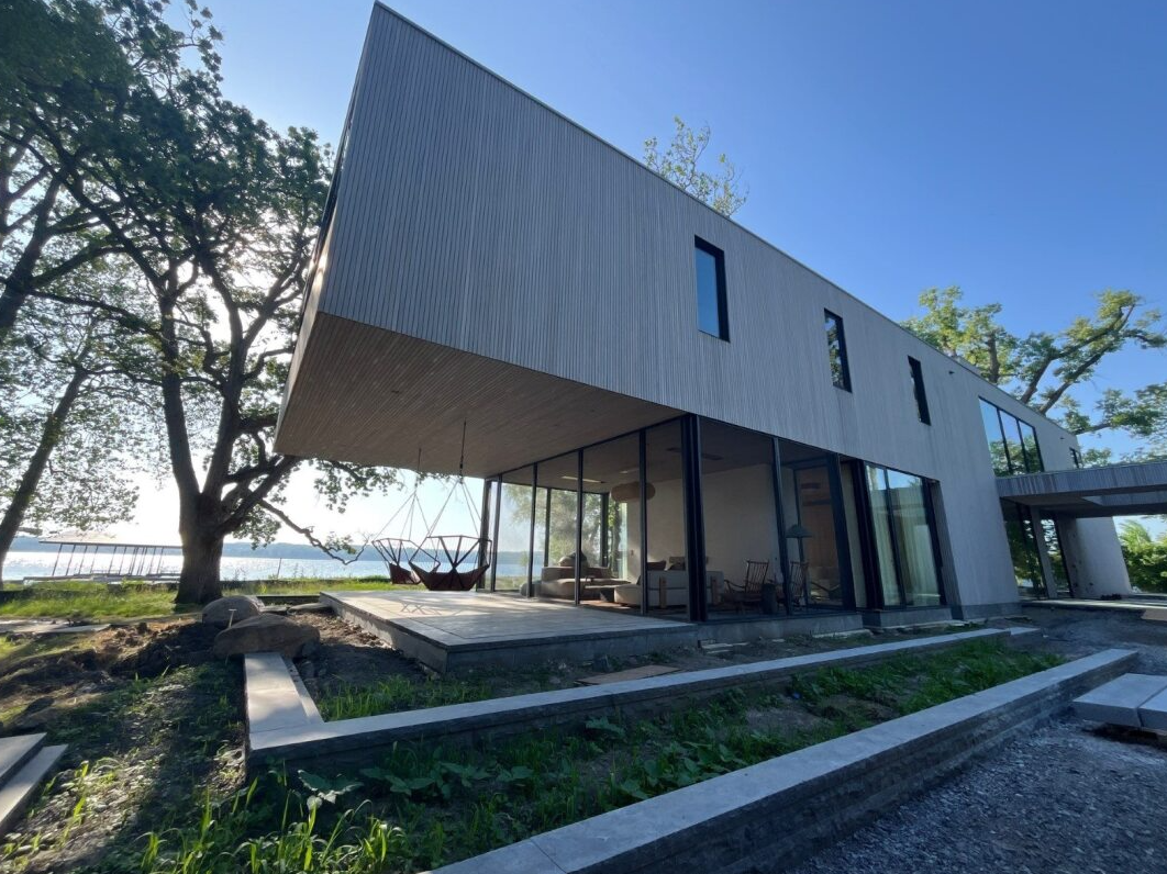 Modern lakeside house with slanted roof and large windows, surrounded by trees and a grassy yard.