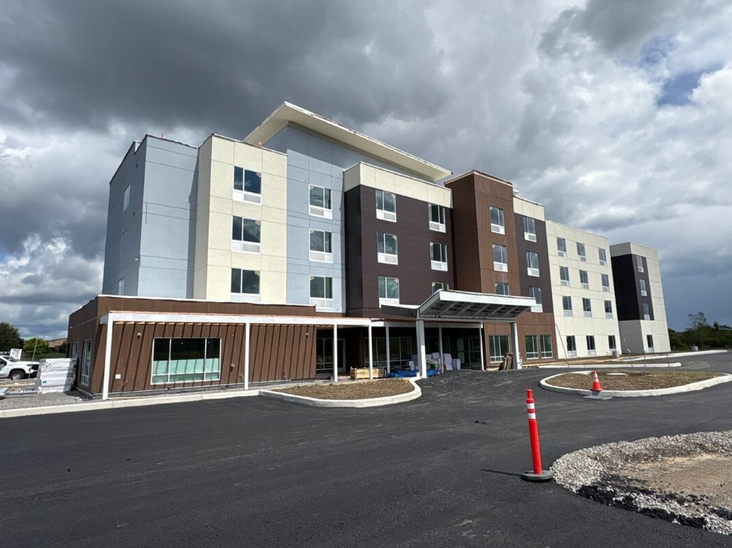 Modern multi-story building with gray and brown exterior under a cloudy sky, viewed from a parking lot.