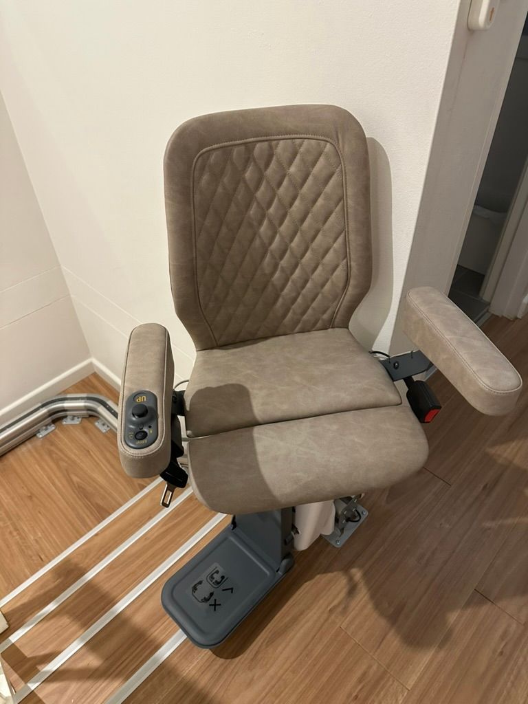 Beige motorized stair lift installed on wooden stairs, armrests and seat.