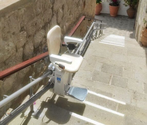 Outdoor stairlift with white seat on a narrow stone stairway beside a wall and handrails, in bright sunlight