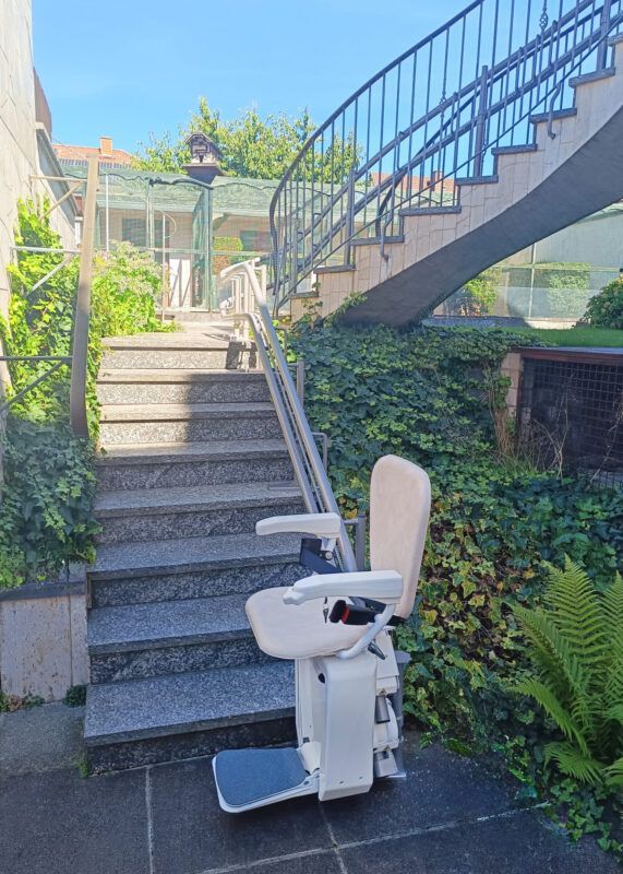 Wheelchair-style stair lift beside outdoor stone steps under a metal railing, surrounded by greenery.
