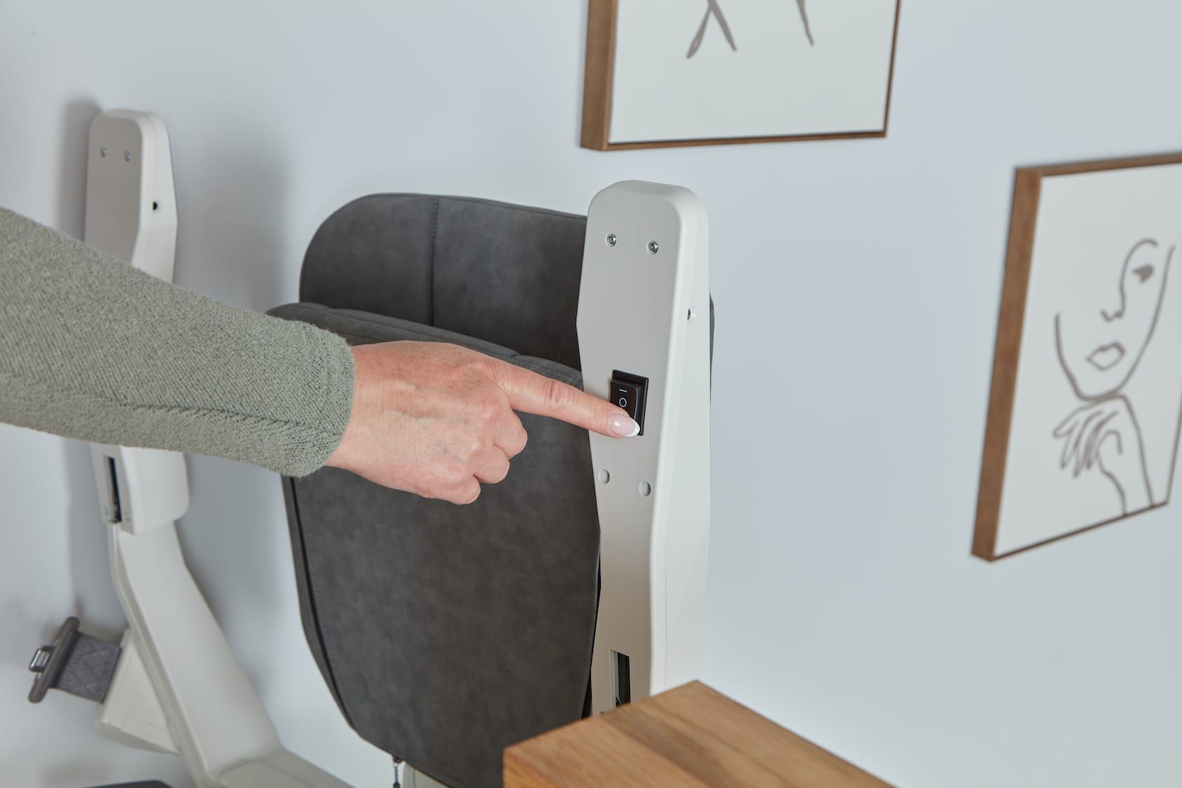 Hand pointing at a white wall-mounted device beside a gray office chair and framed sketches