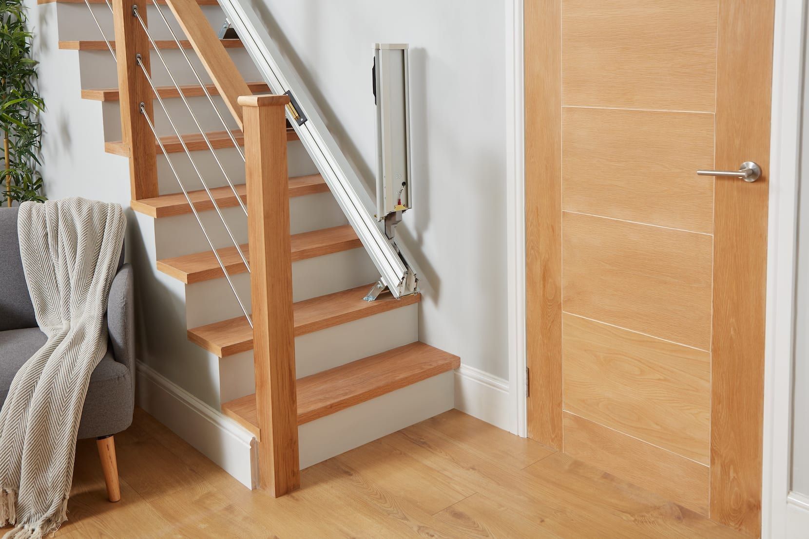 Modern wooden staircase beside a closed door in a bright hallway, with a chair and blanket on the left