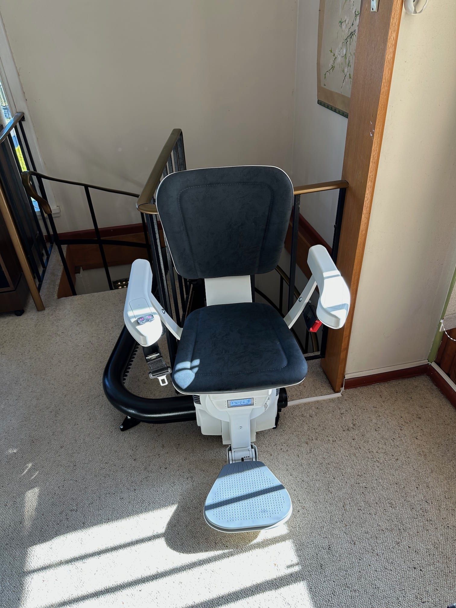 White and blue stair lift chair on carpeted stairs by a doorway, with sunlight across the floor.