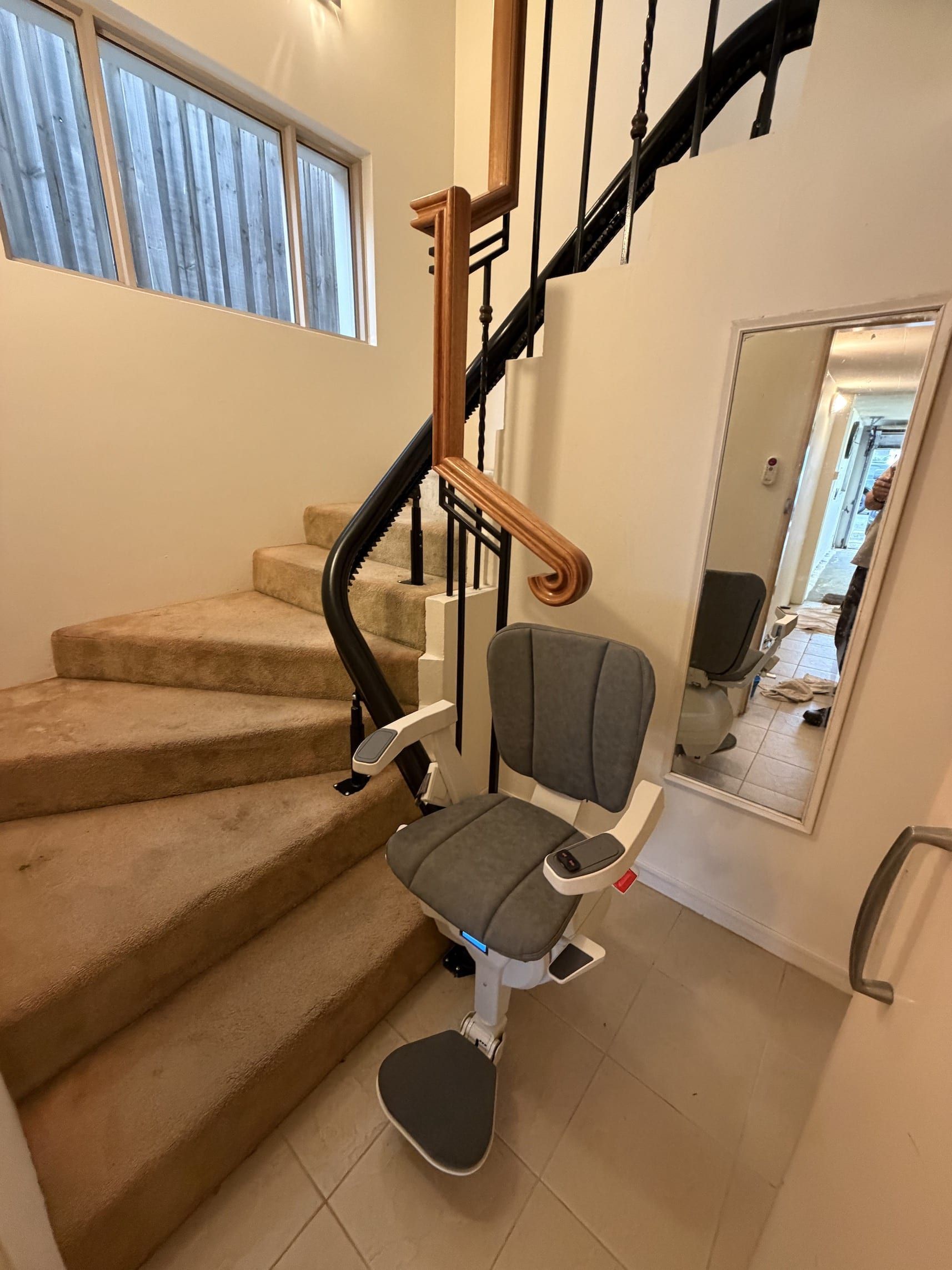 Stairlift chair on a carpeted staircase in a home interior, with beige walls and a hallway mirror.