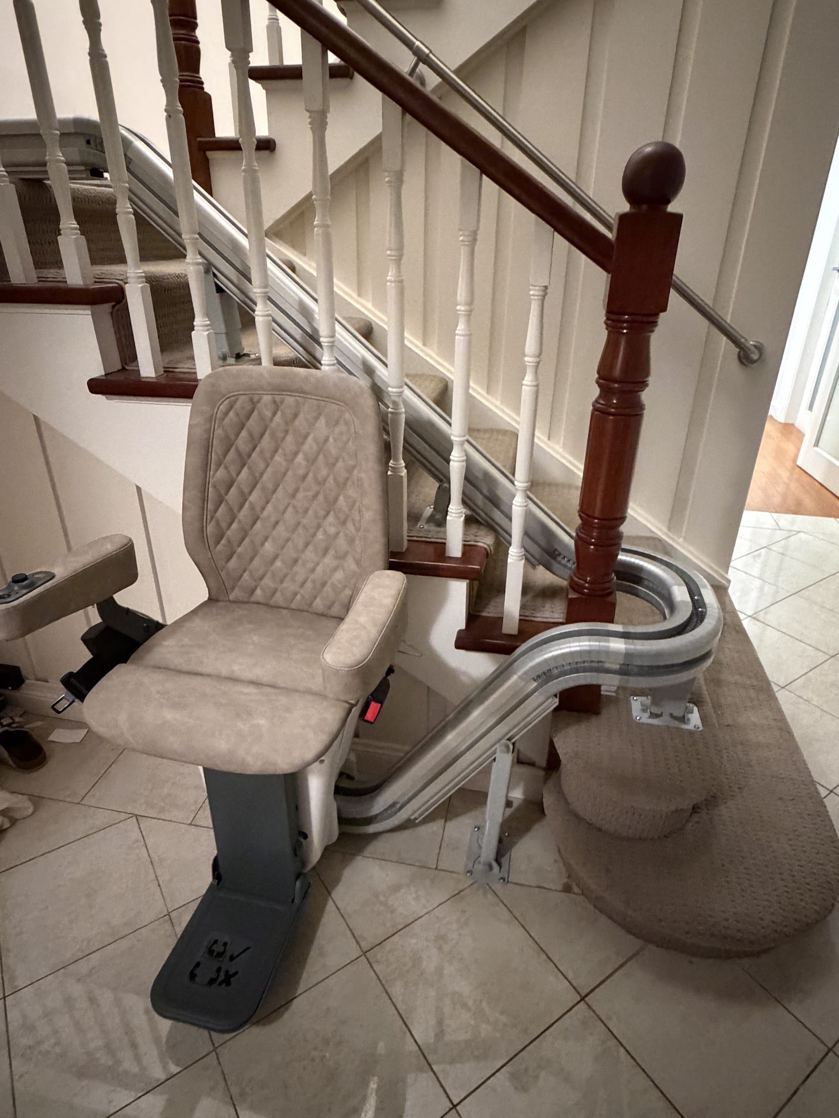 Curved stairlift installed on a carpeted staircase beside a beige upholstered seat and tiled entryway