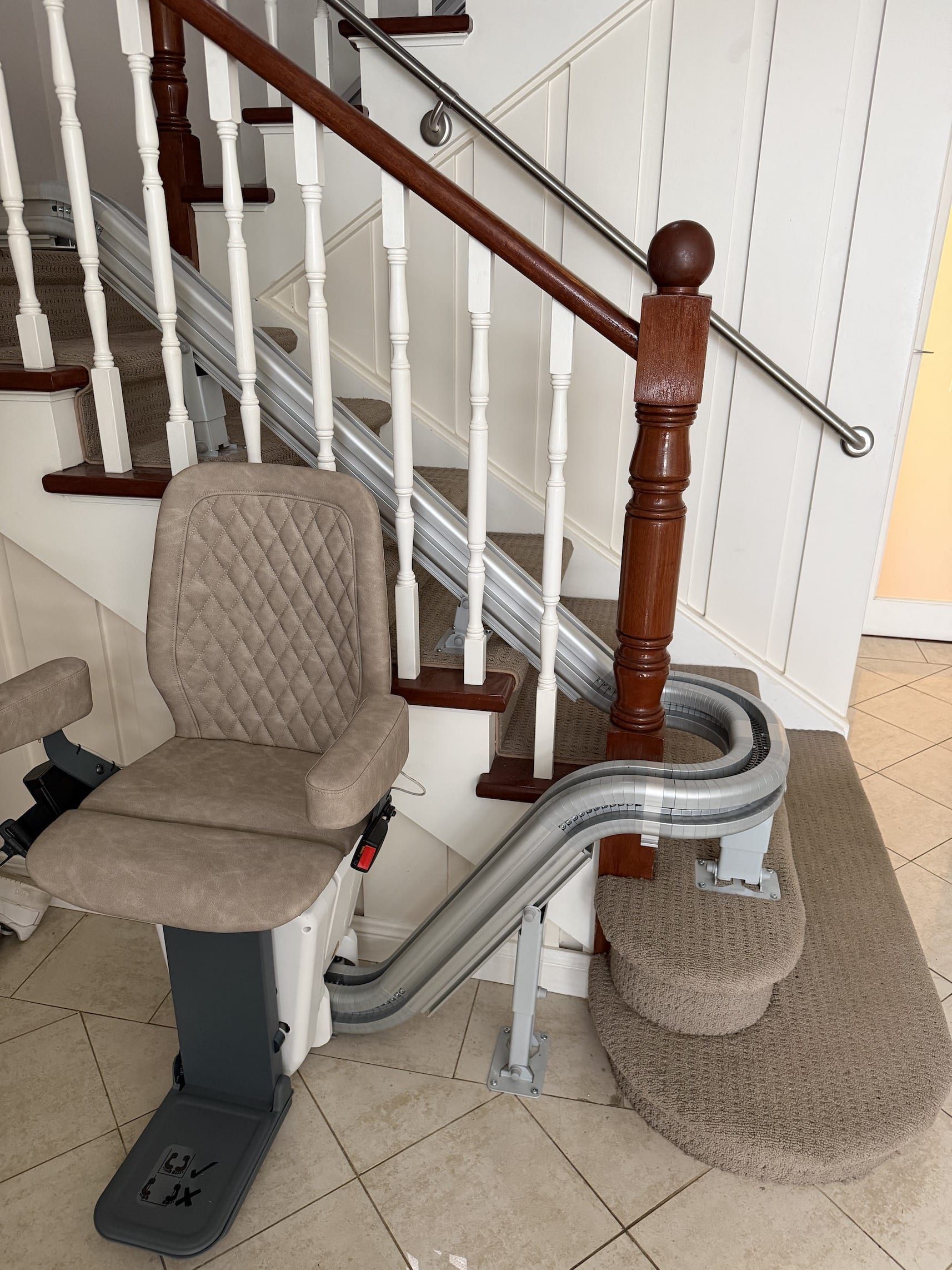 Stair chair lift beside a carpeted staircase with white banisters and a beige chair seat