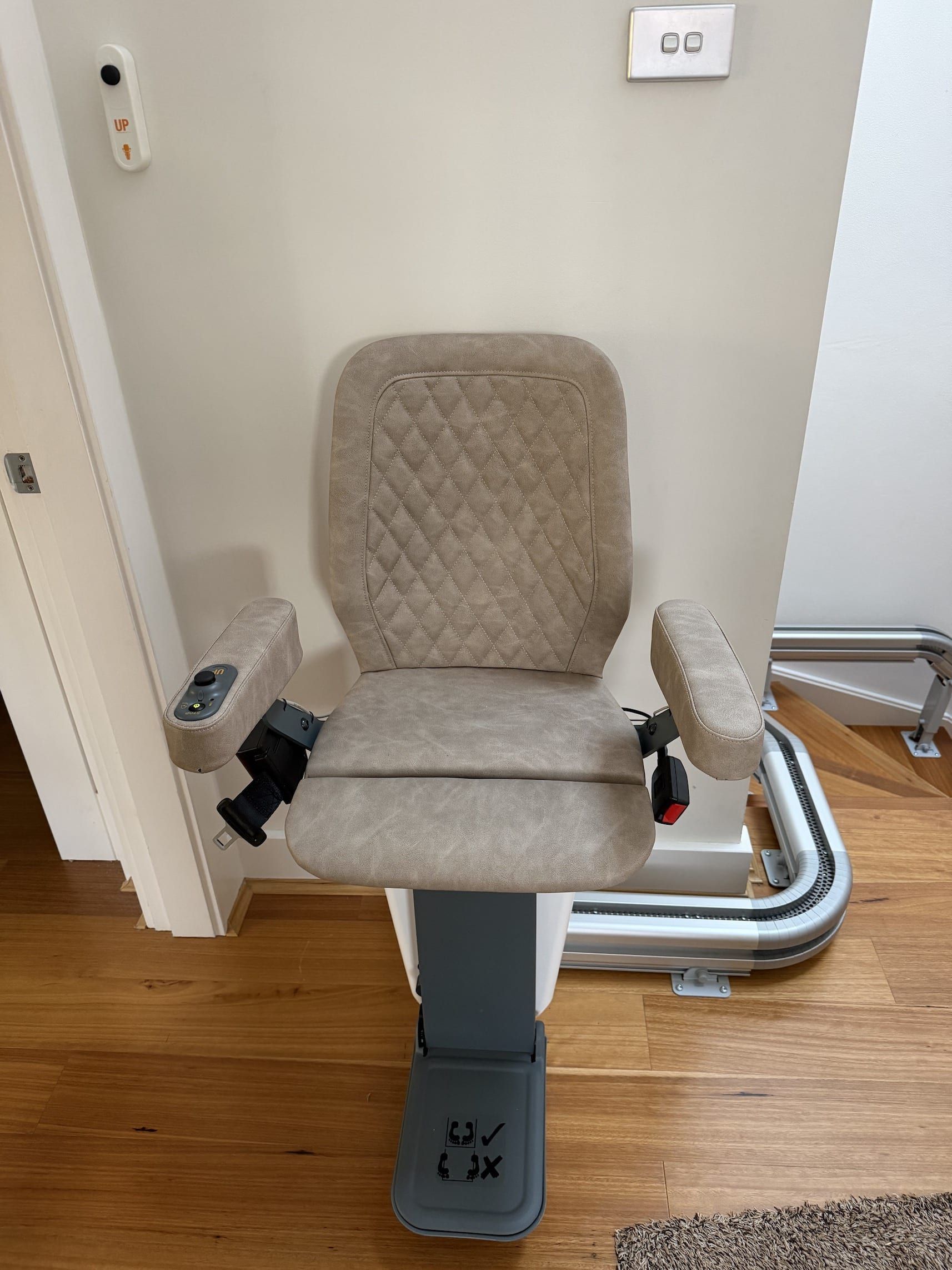 Gray patient lift chair with padded armrests in a room with wood flooring and wall outlets