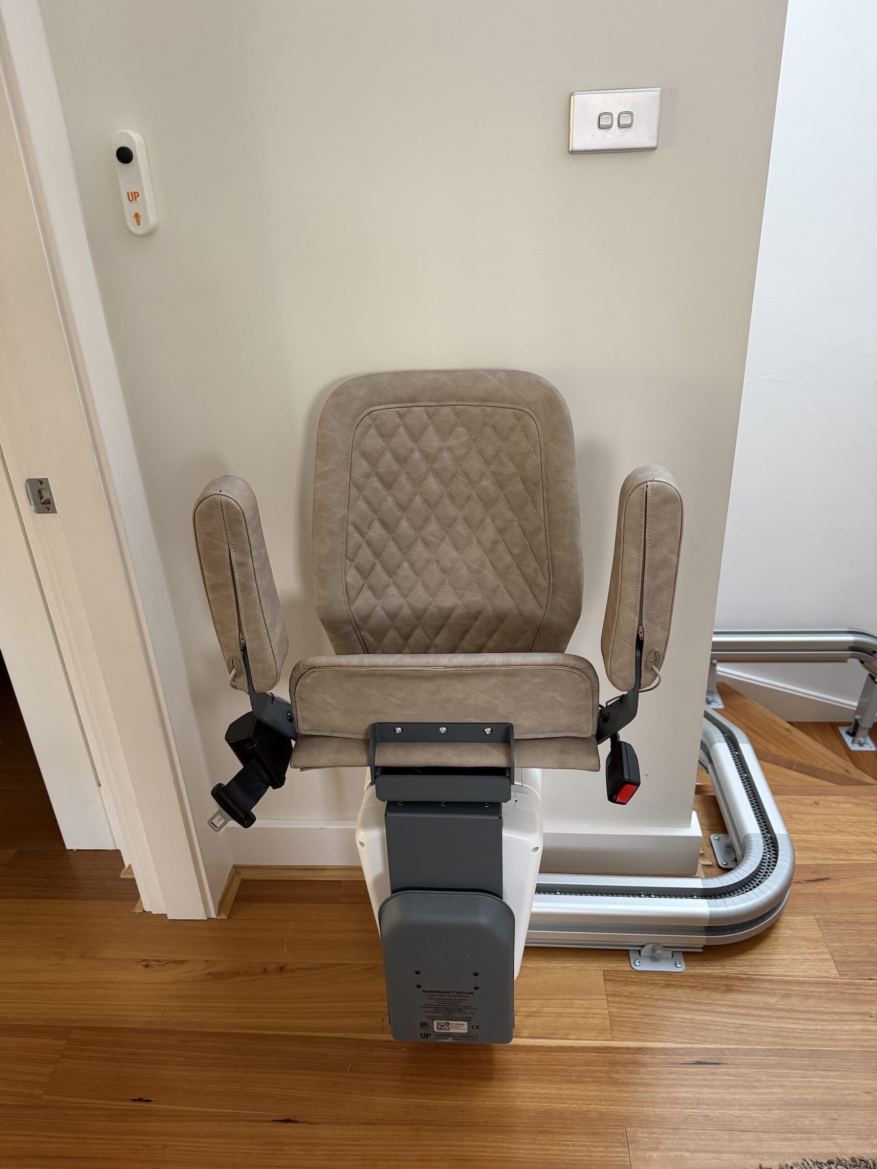 Brown stairlift chair installed on a narrow staircase beside a beige wall and doorway.