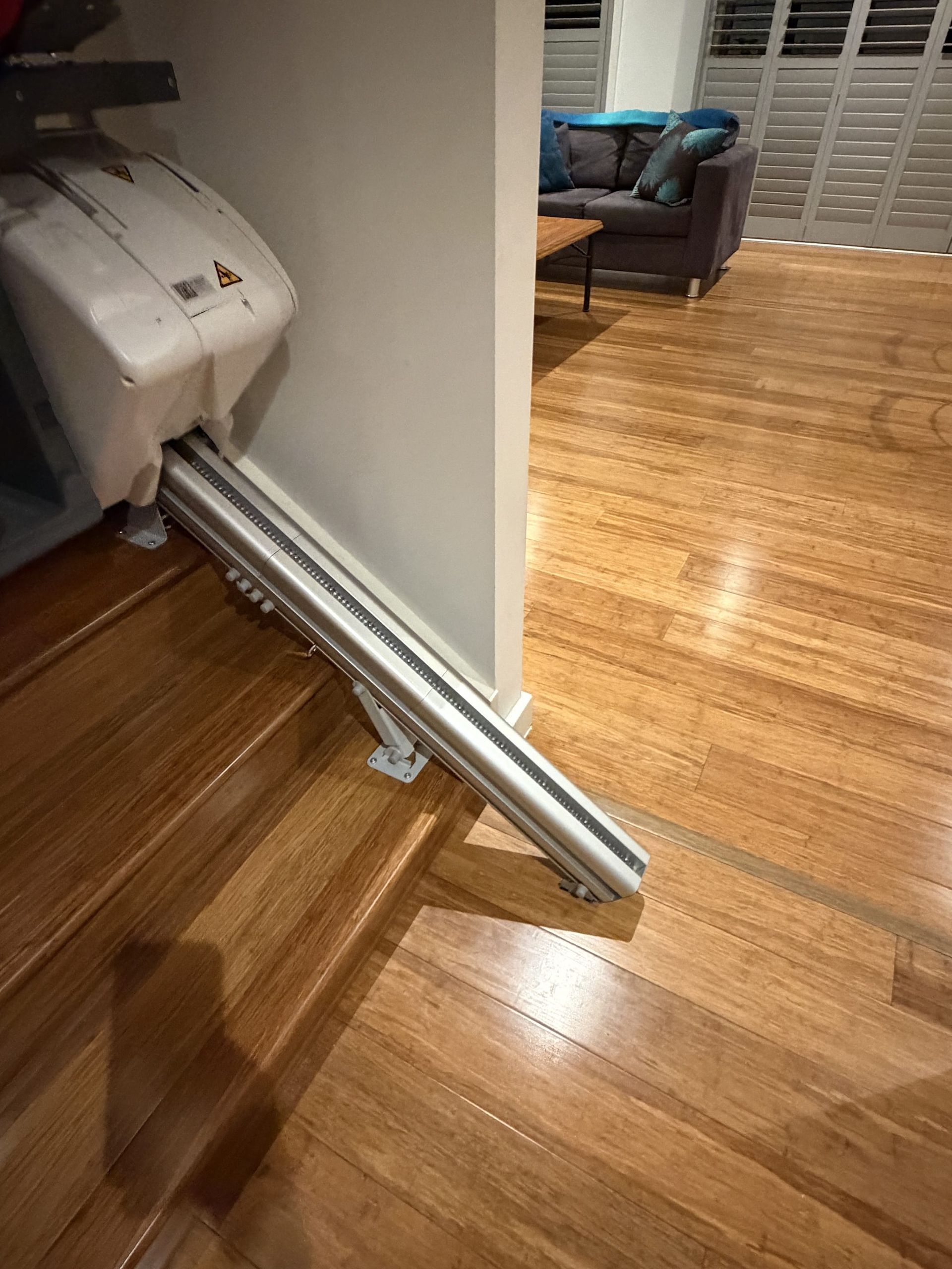 White wheelchair or walker folded against a wall in a hardwood-floored home hallway, leading into a living room.