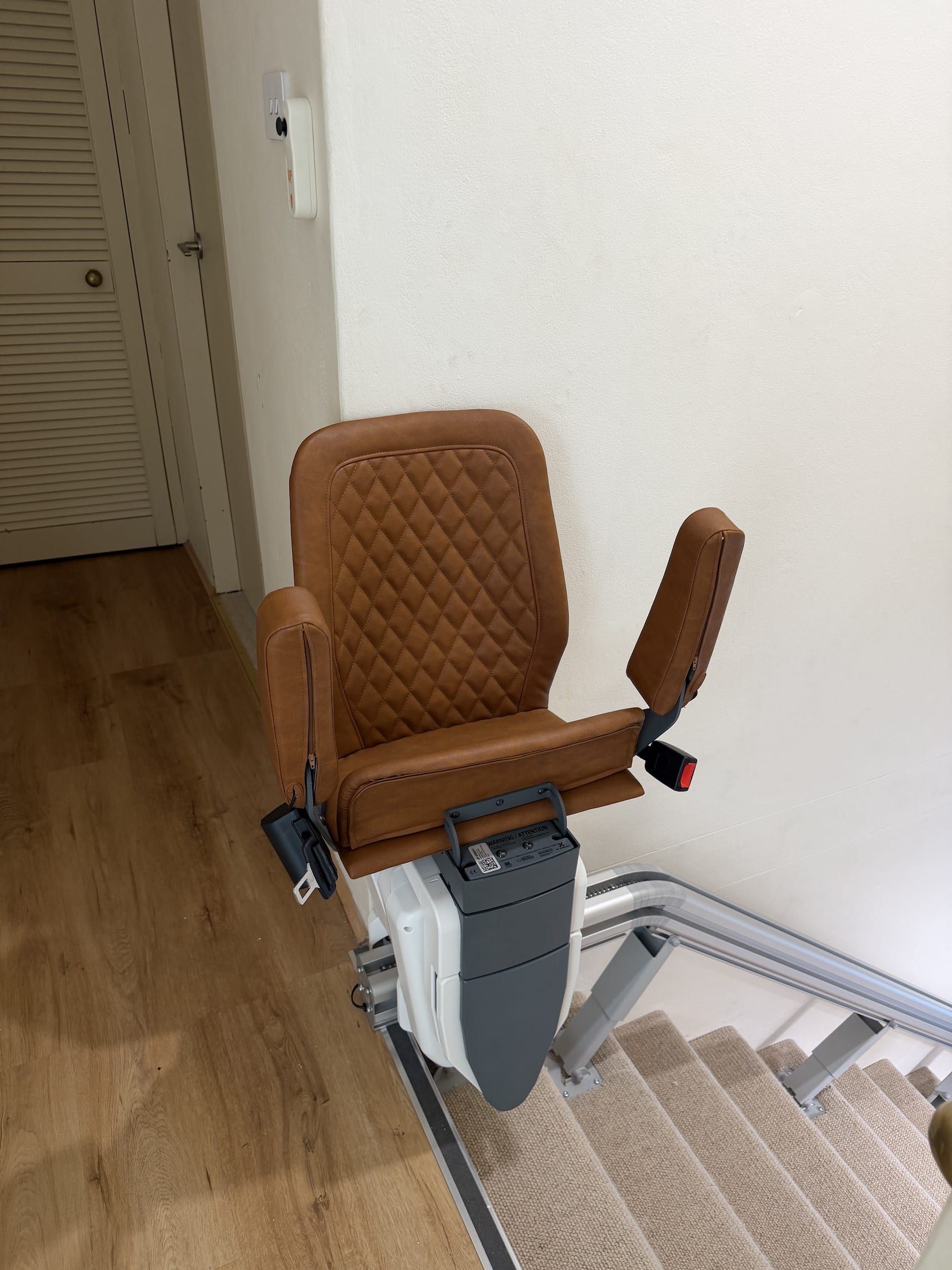 Brown stairlift chair beside a carpeted staircase in a hallway with wood flooring