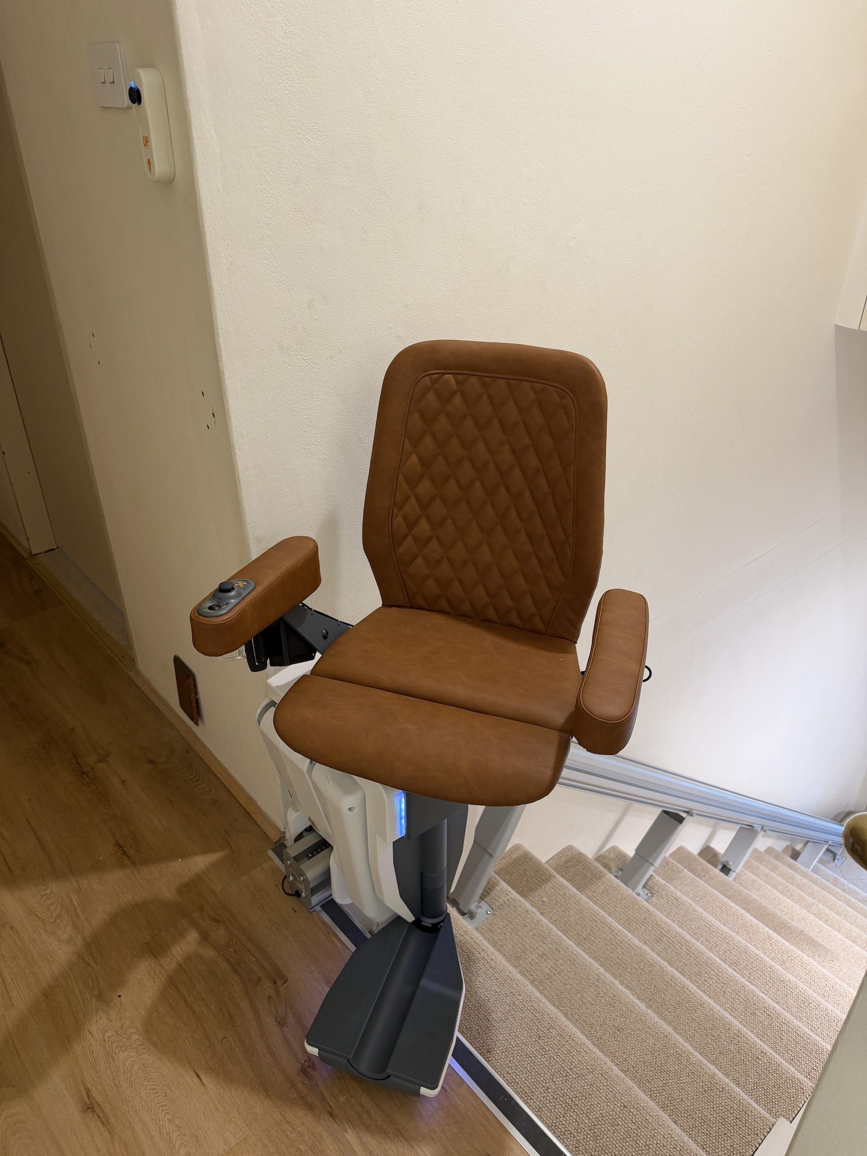 Brown stair lift chair on a carpeted staircase beside a wall and hallway.