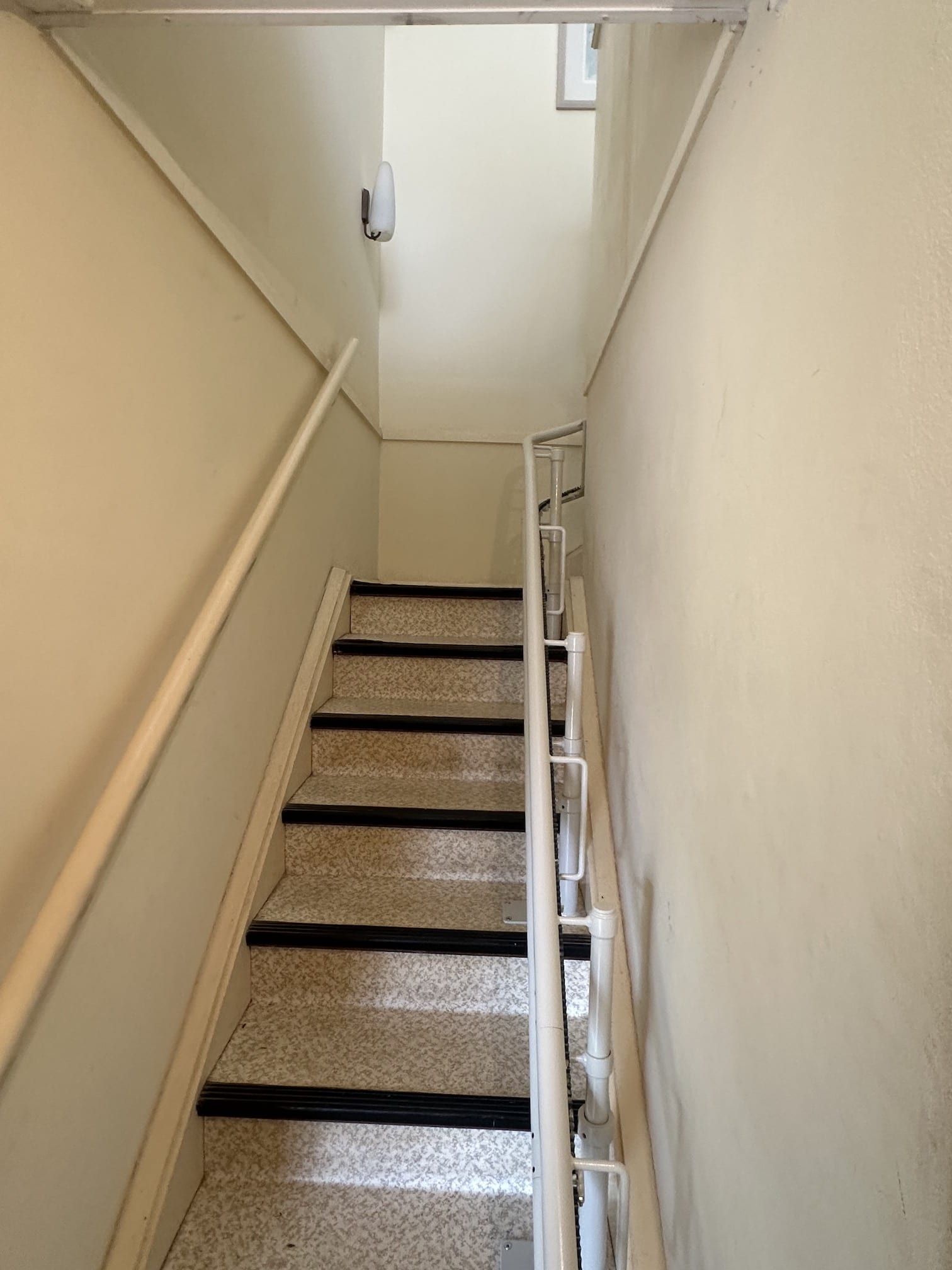 Narrow indoor stairway with beige walls, carpeted steps, and a handrail along the right side
