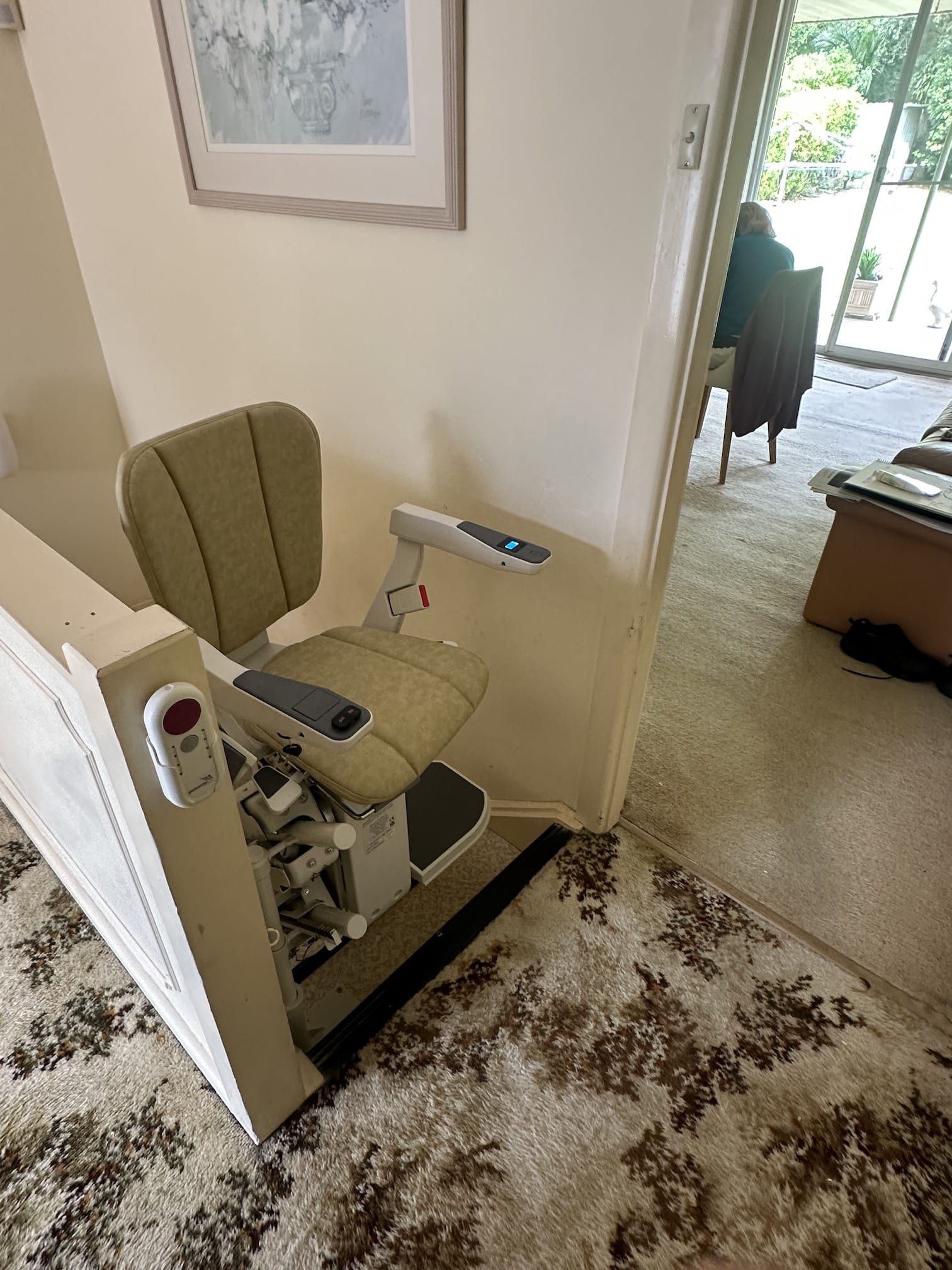 Stairlift chair beside a carpeted hallway, facing an open doorway to a sunlit room.