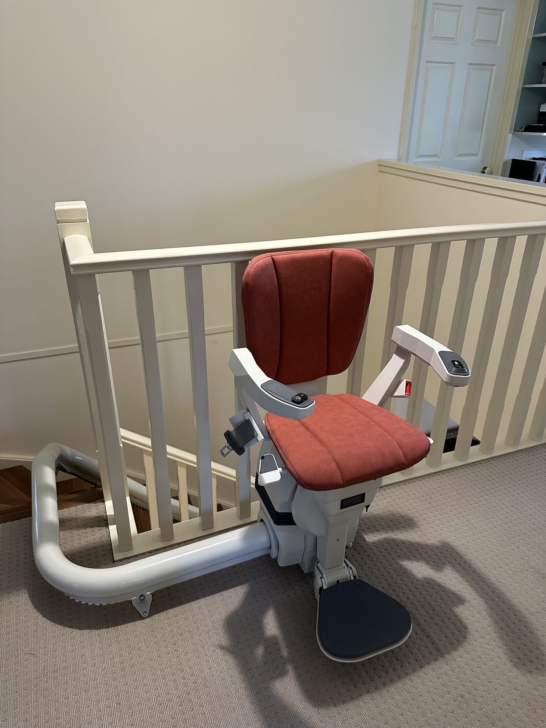 Red stair lift chair with gray frame beside a white banister on a carpeted landing.