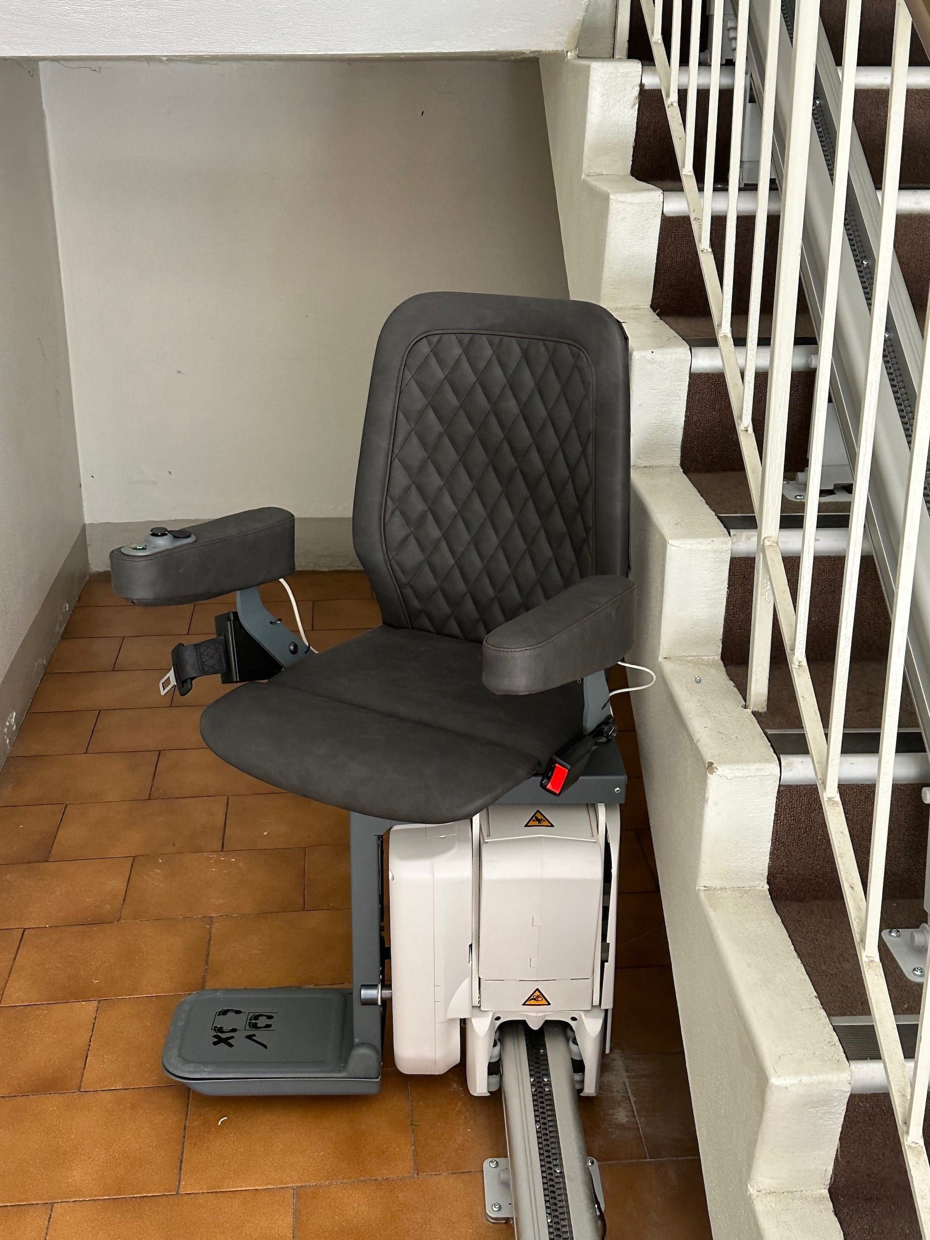 Stairlift chair beside indoor stairs, with a gray seat and quilted backrest.