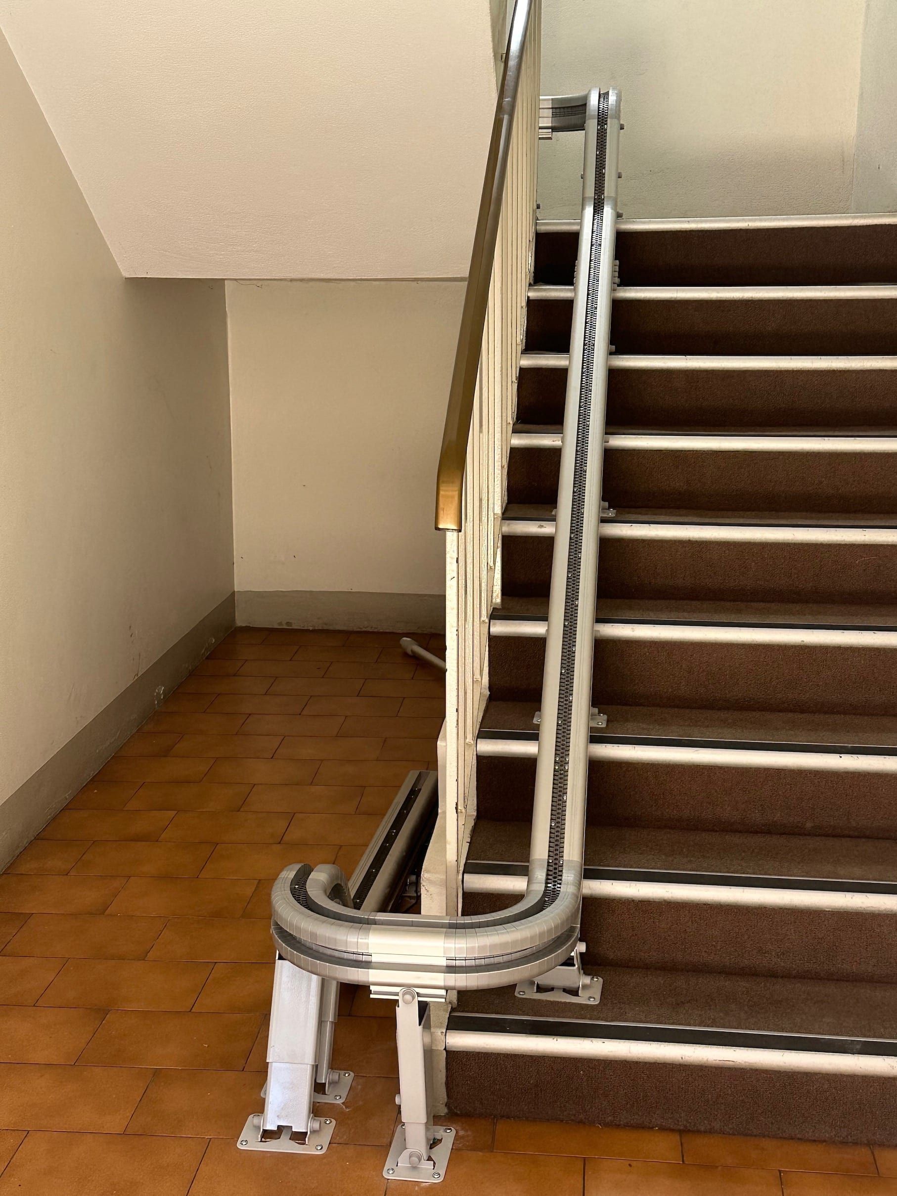 Indoor stairwell with a metal stairlift rail along carpeted stairs and a tiled landing below