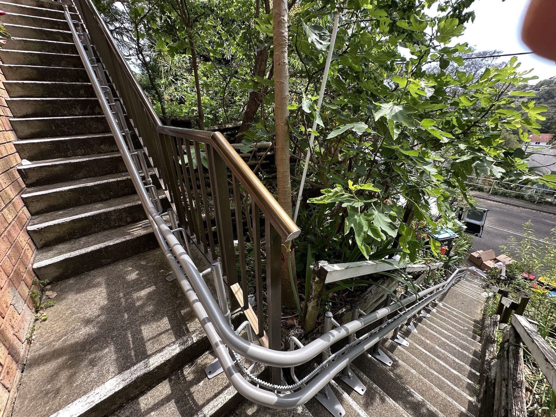 Steep outdoor stairway with metal railings and trees beside a brick wall and road