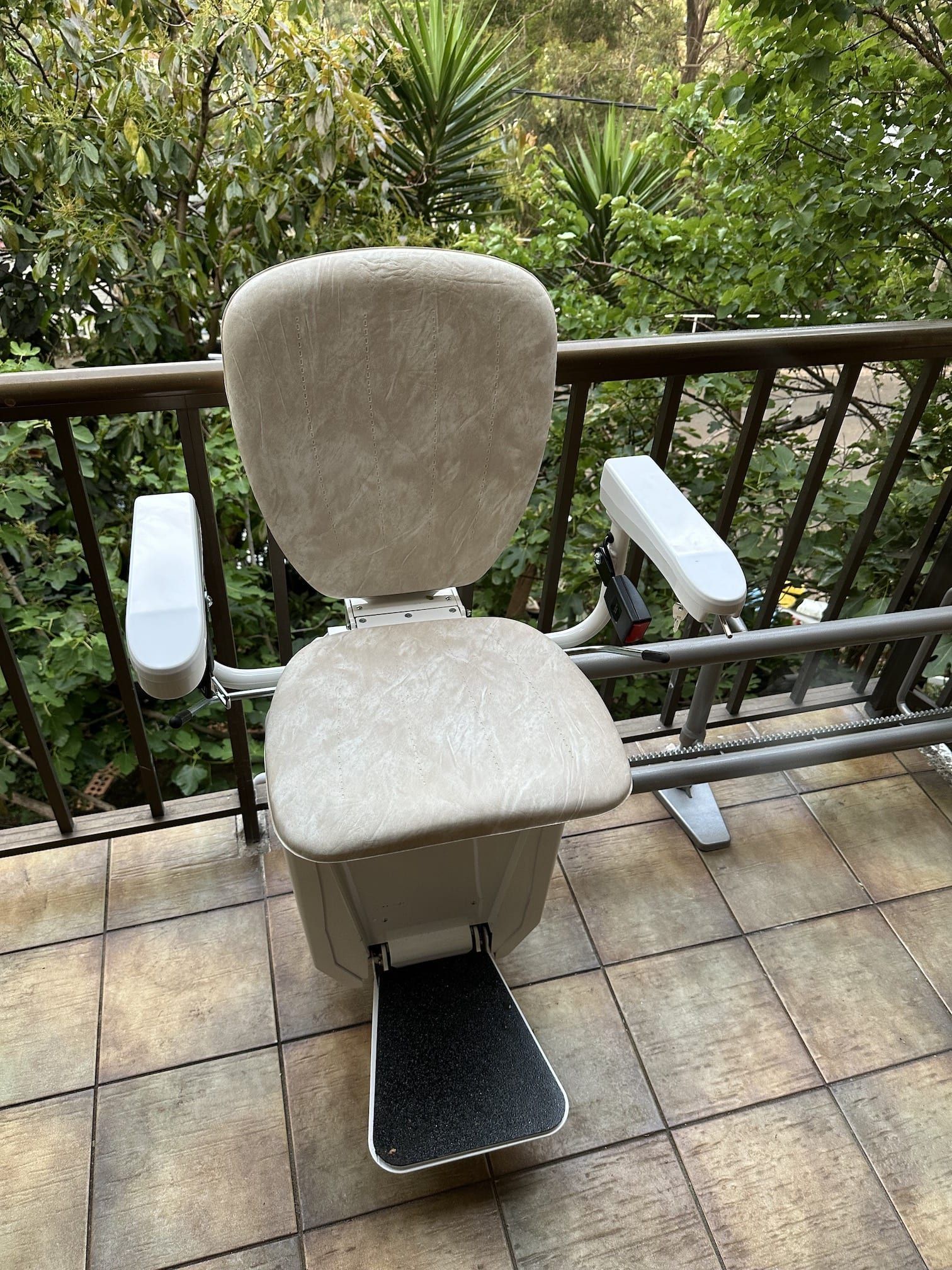Outdoor stairlift chair with beige seat and footrest on a tiled patio overlooking greenery