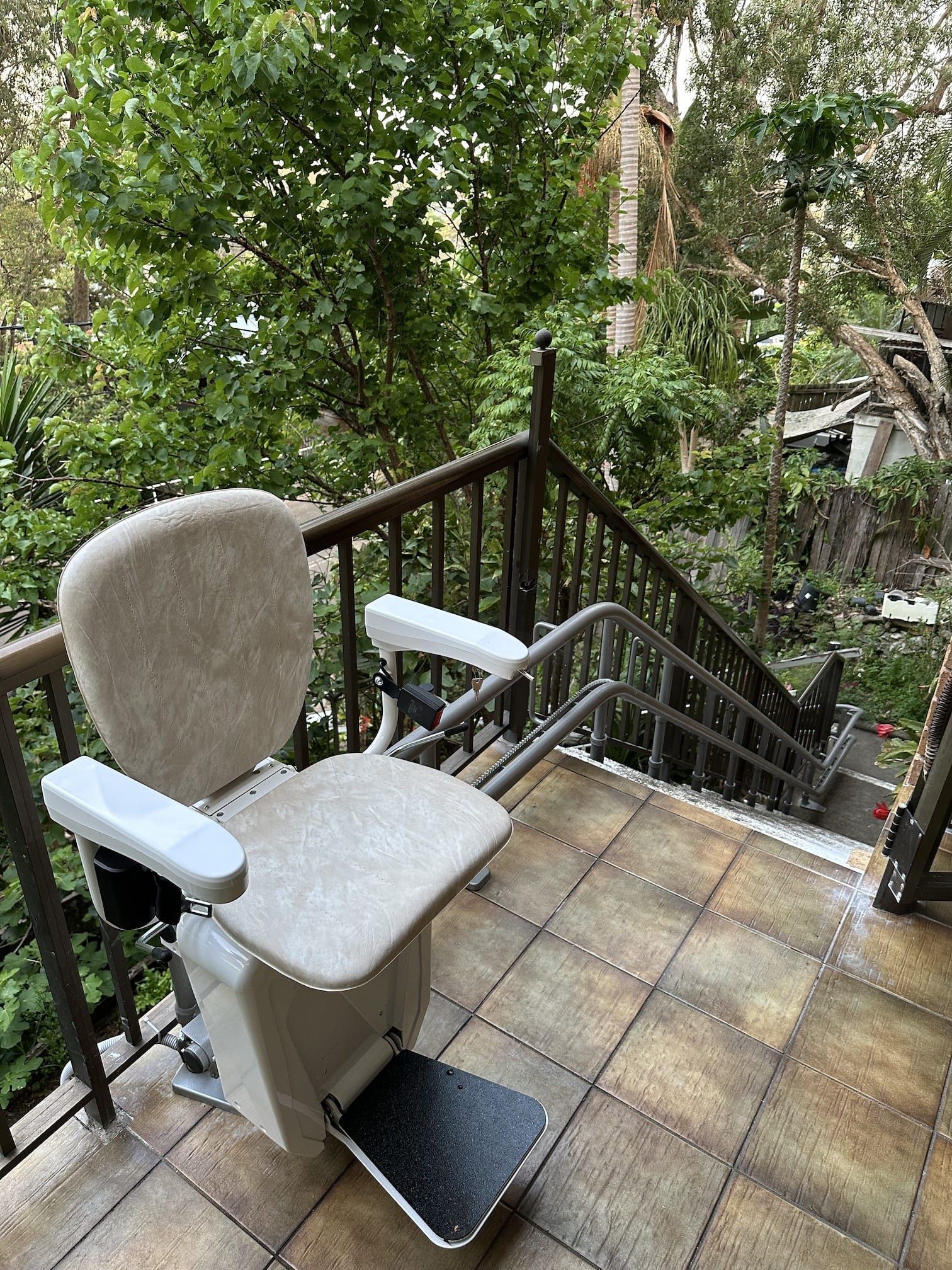 Outdoor stairlift on a tiled patio with a beige seat beside a black railing and lush greenery.