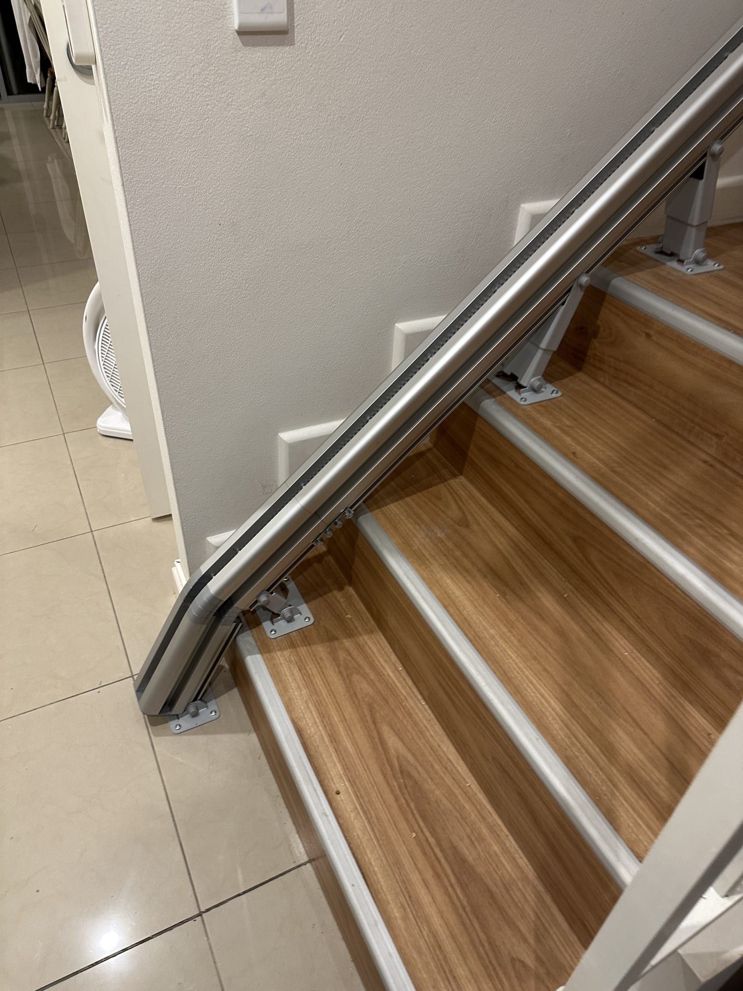 Indoor staircase with wood treads, silver railings, and beige tile floor beside a wall.