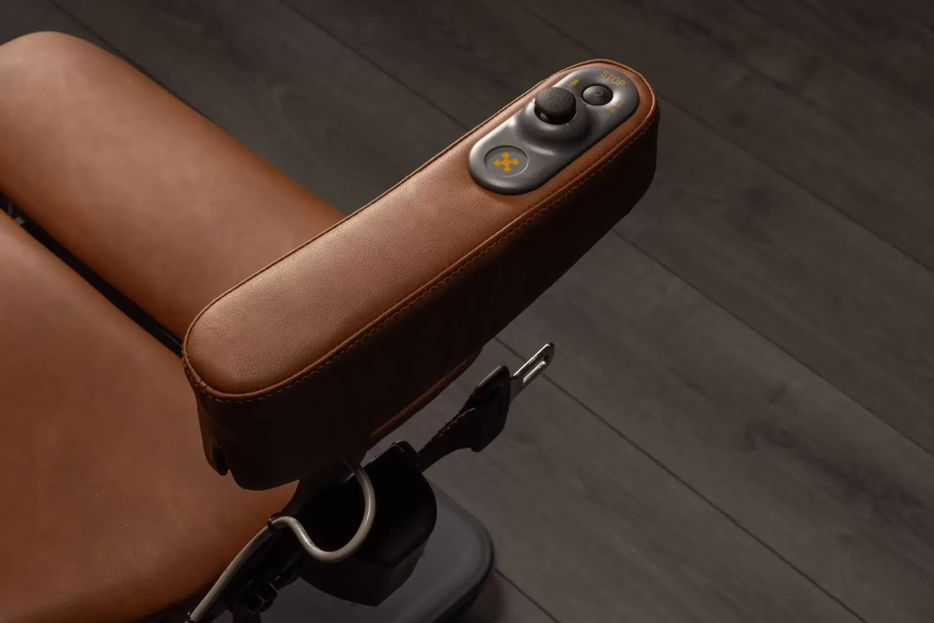 Brown leather wheelchair armrest featuring a central control joystick and buttons, positioned above a dark wood floor.