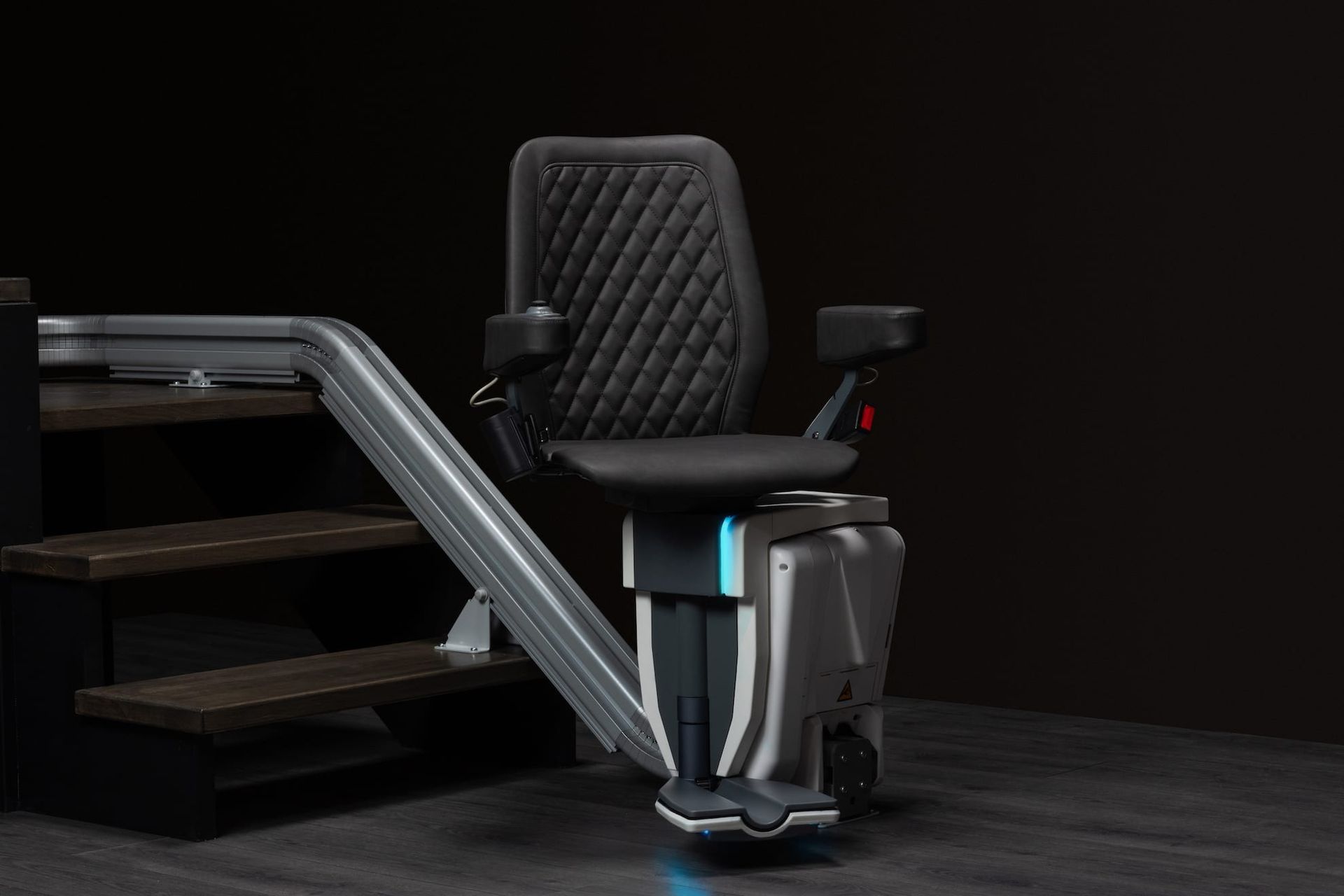 Modern stairlift chair beside carpeted stairs in a dark interior, with black seat and silver rail