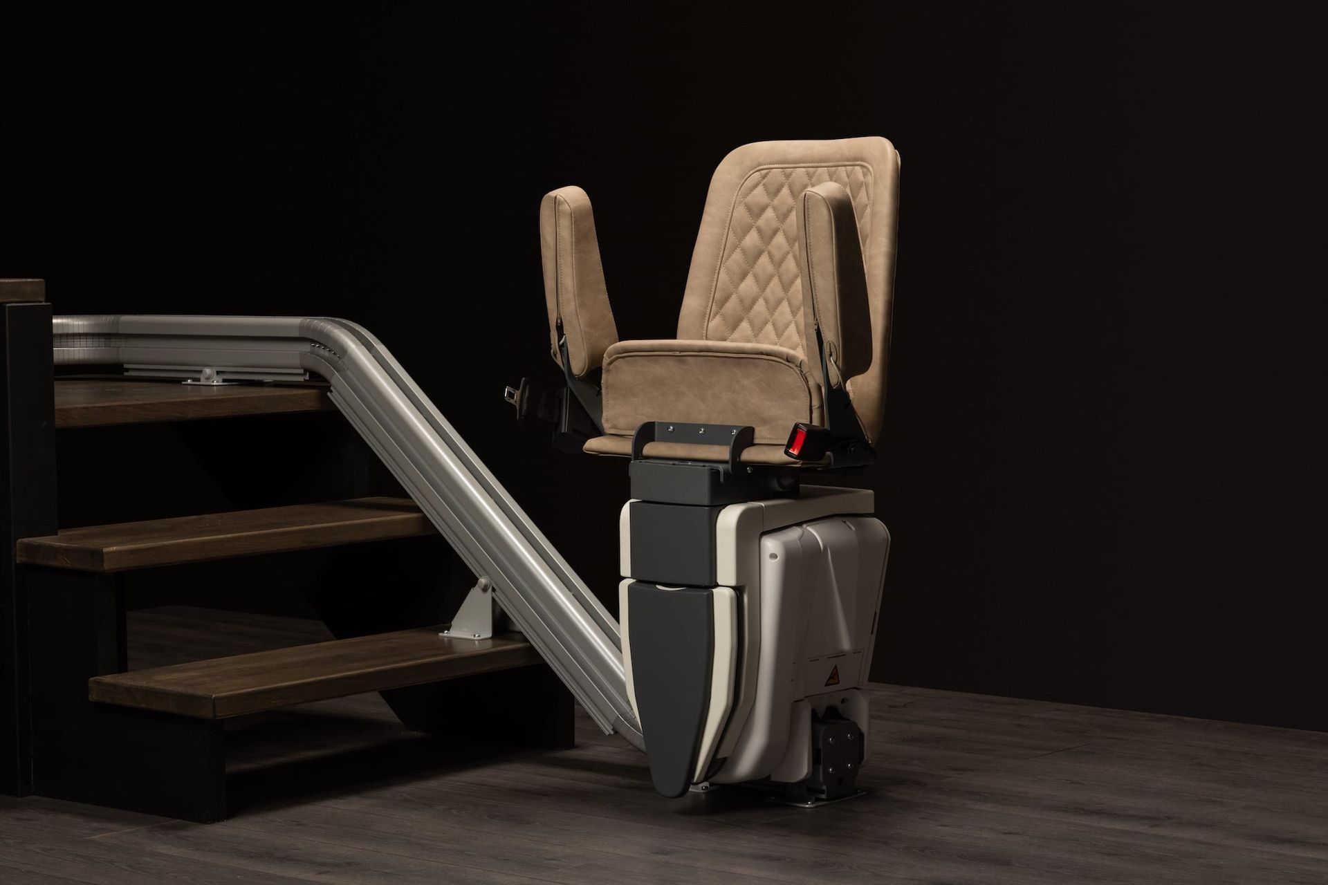 Compact stairlift with beige quilted seat and silver rail on a dark stairway.