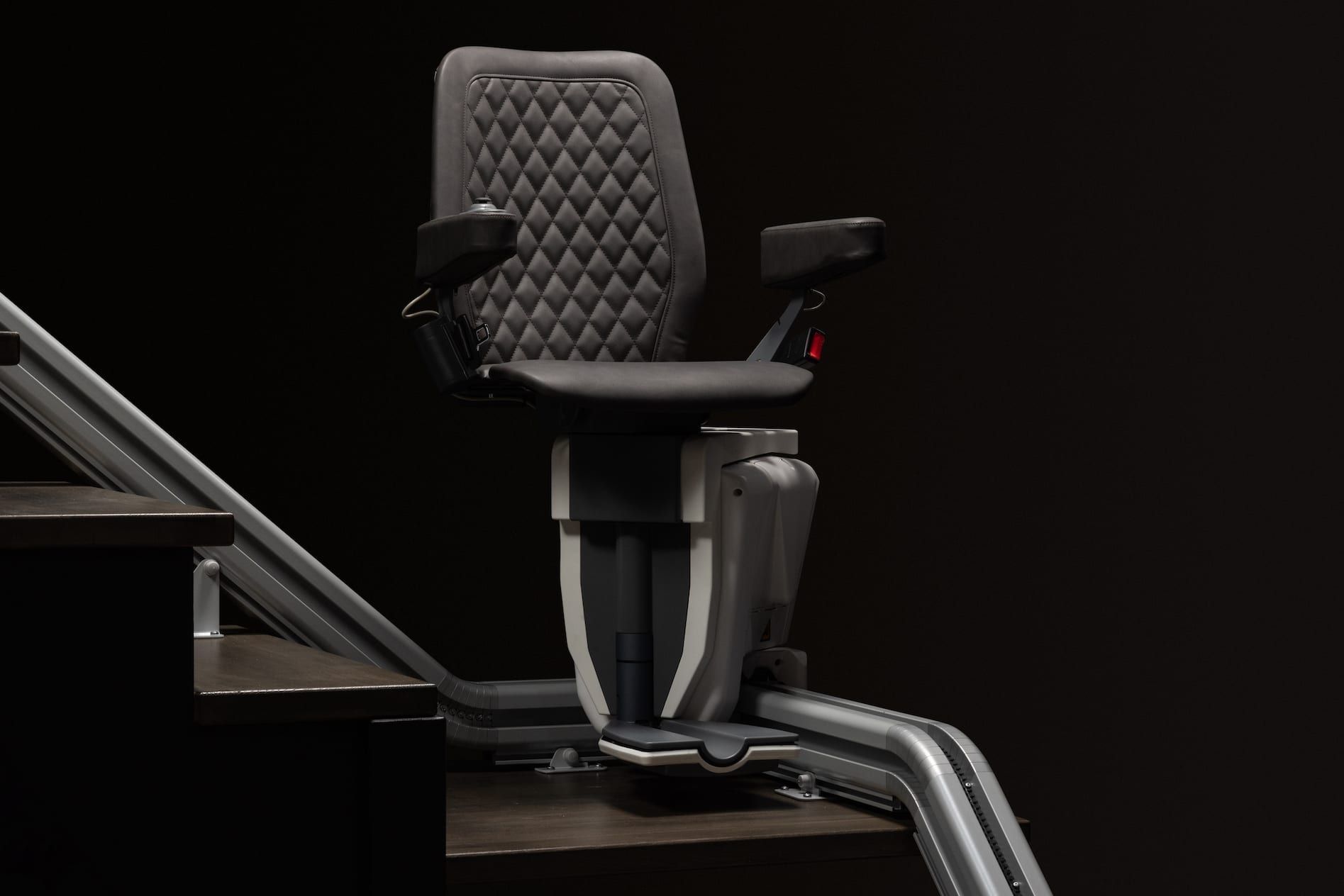 Gray stairlift chair on a dark staircase, with metal rails and padded seat against a black background