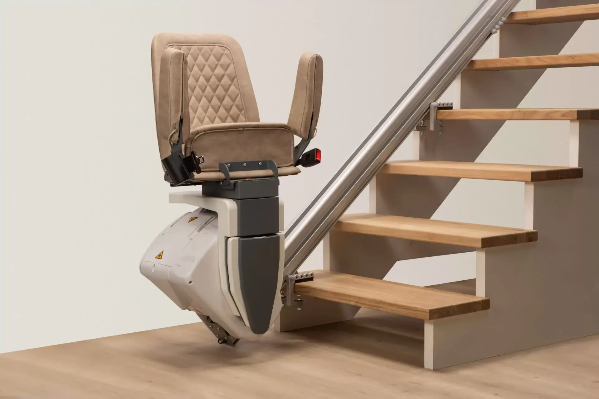 A beige upholstered stairlift positioned at the bottom of a wooden staircase, mounted on a metal rail.