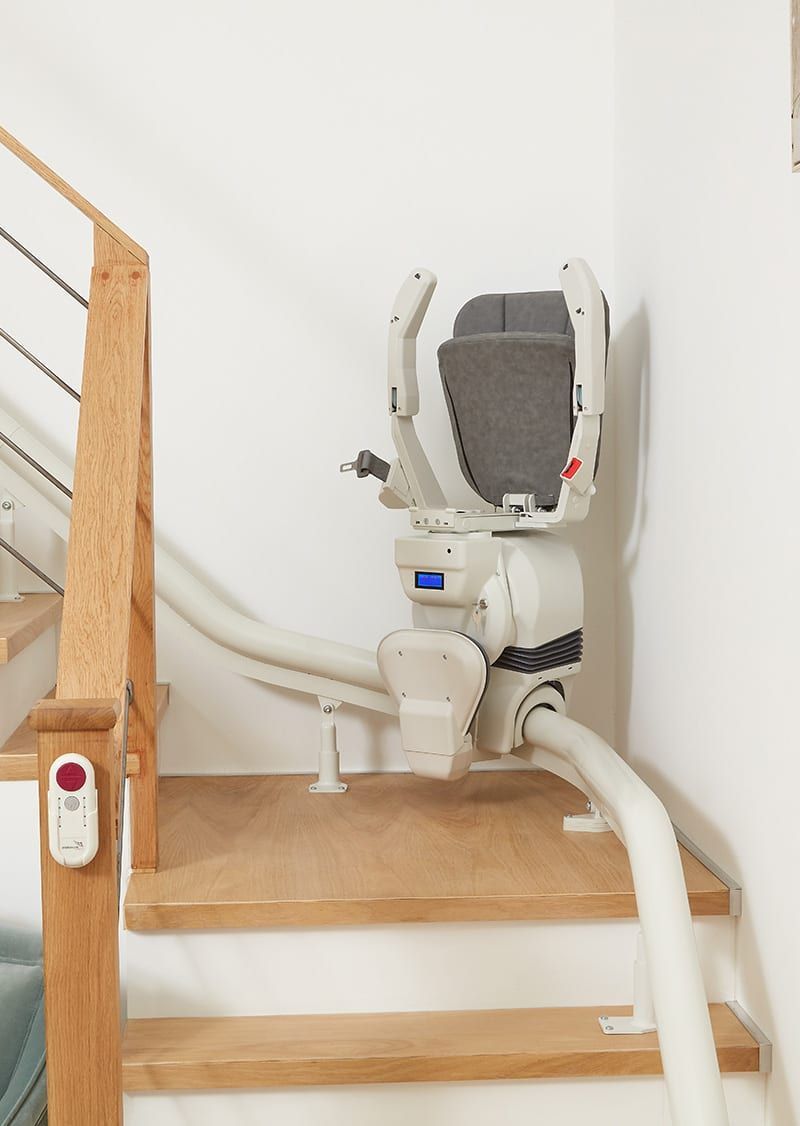 Stairlift on wooden stairs with white walls. The chair is unoccupied, angled towards the top