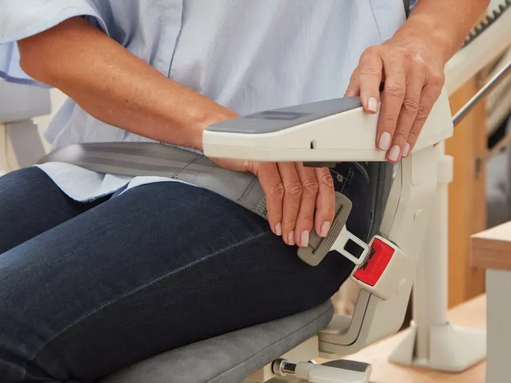 A person sitting in a stairlift, wearing a grey seatbelt secured by a red buckle.