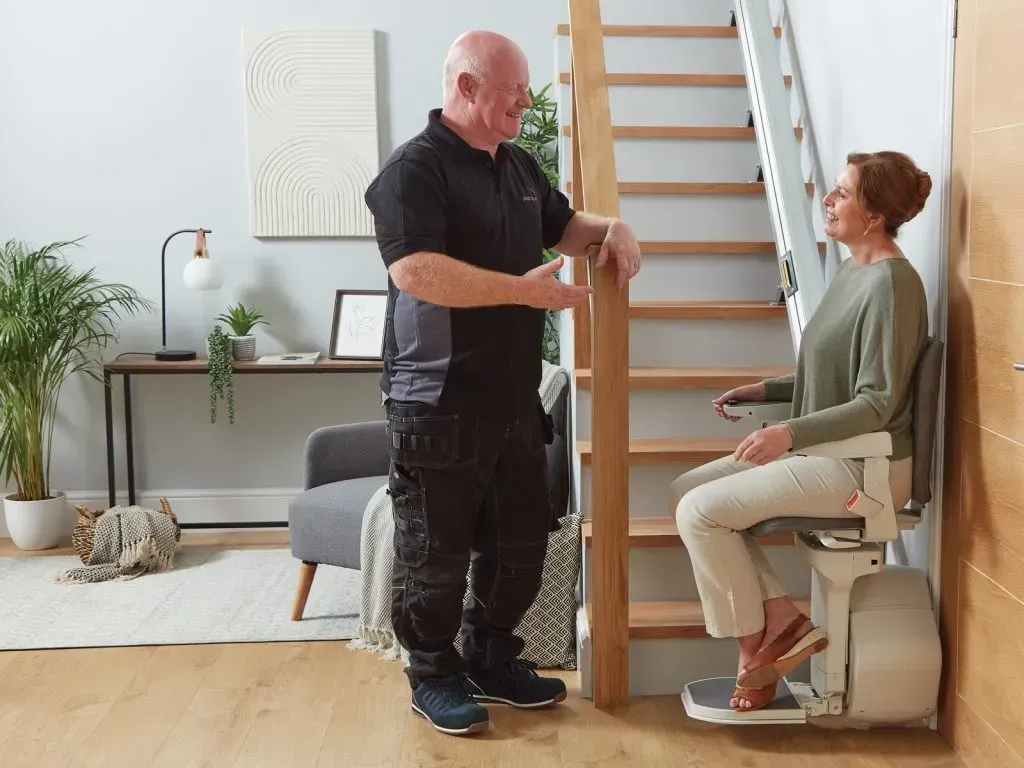 A smiling person sitting on a beige stairlift installed next to wooden stairs inside a home.
