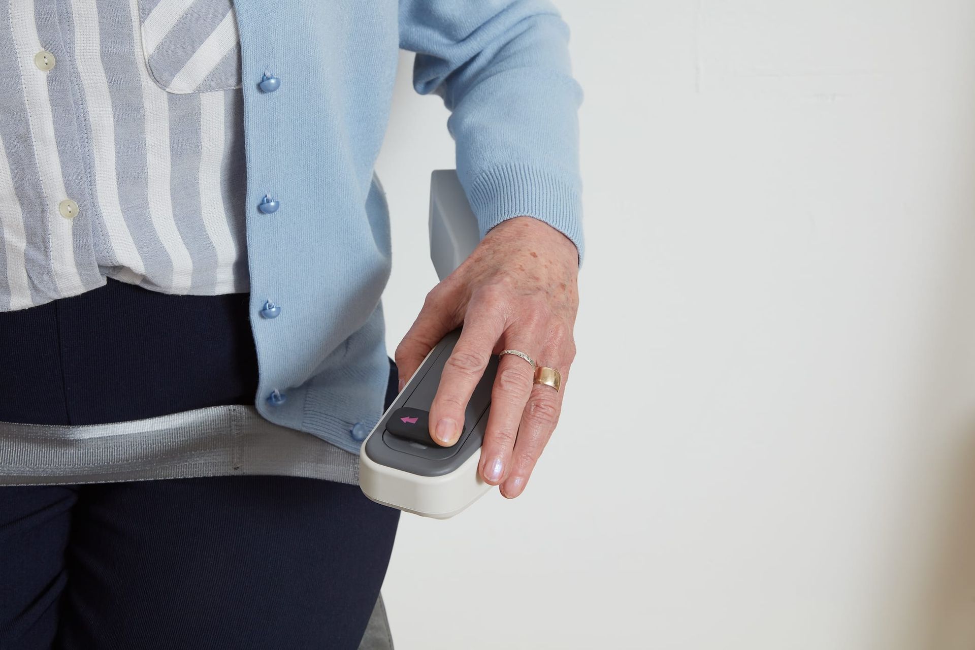 Person's hand operating a button on a chair lift armrest. Blue sweater, striped shirt, white wall.