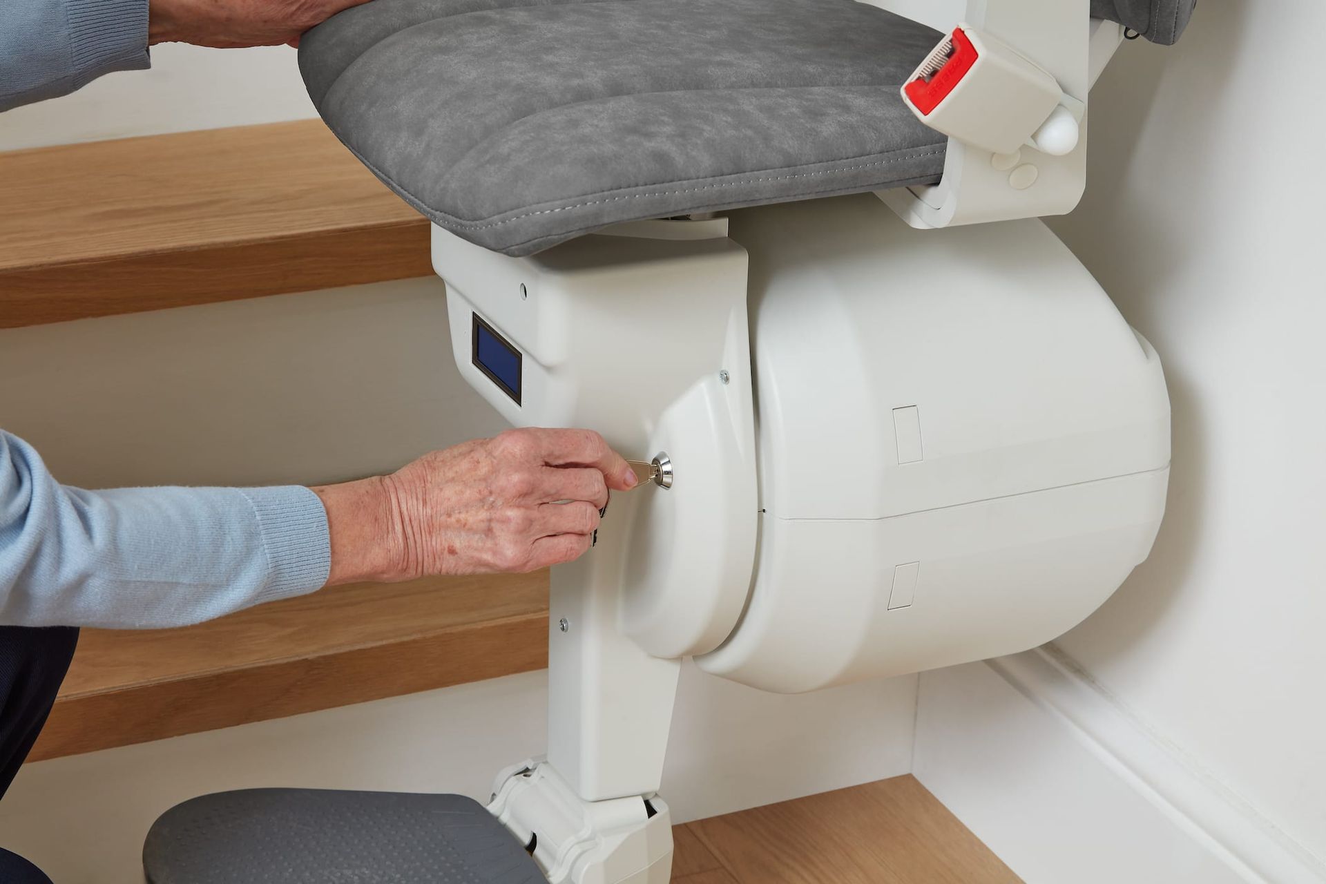 Person using key to turn on a stairlift. Grey seat and cream-colored lift. Wooden stairs.