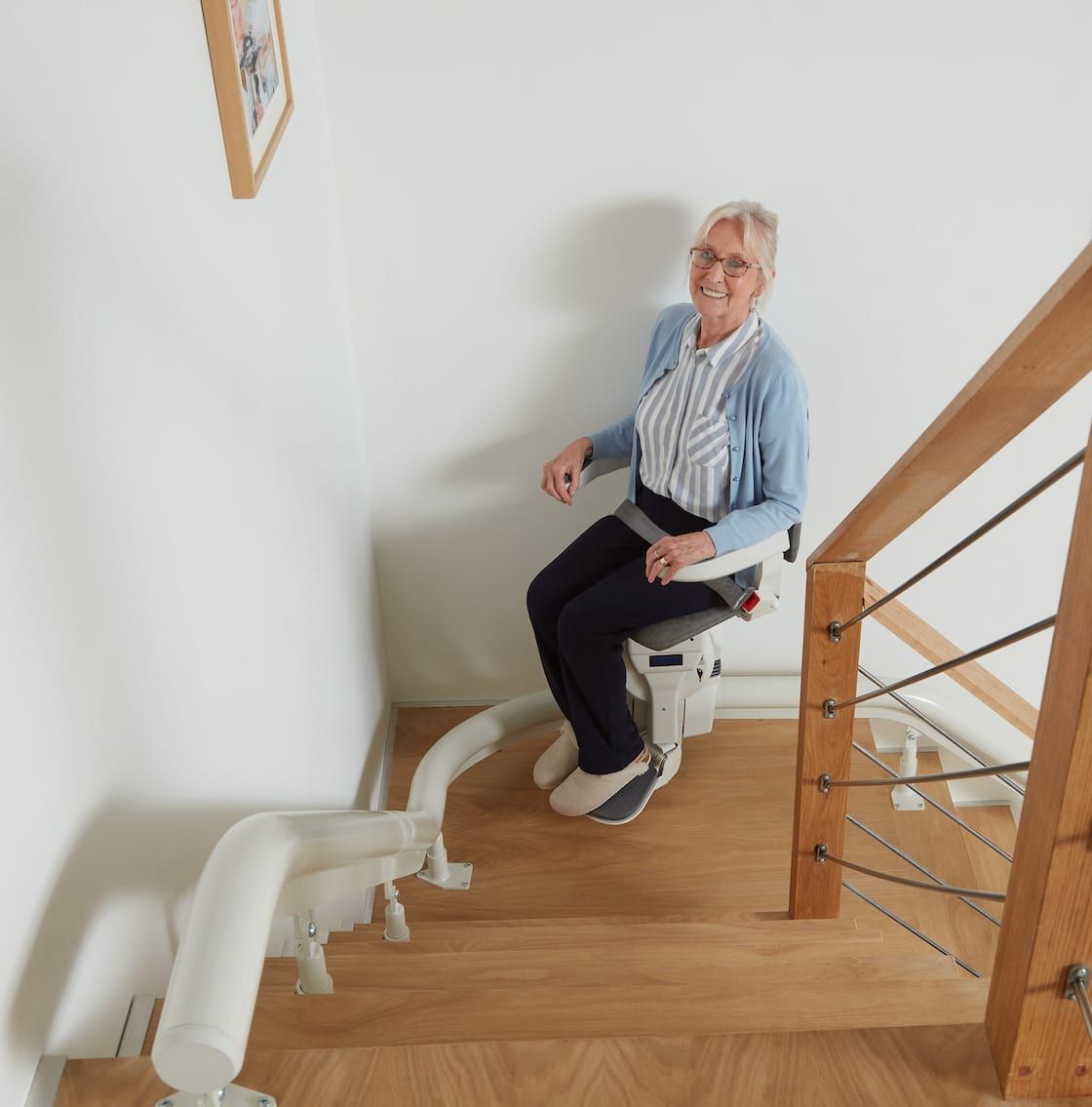 Woman on a stairlift smiles, ascending a wooden staircase beside a white wall.