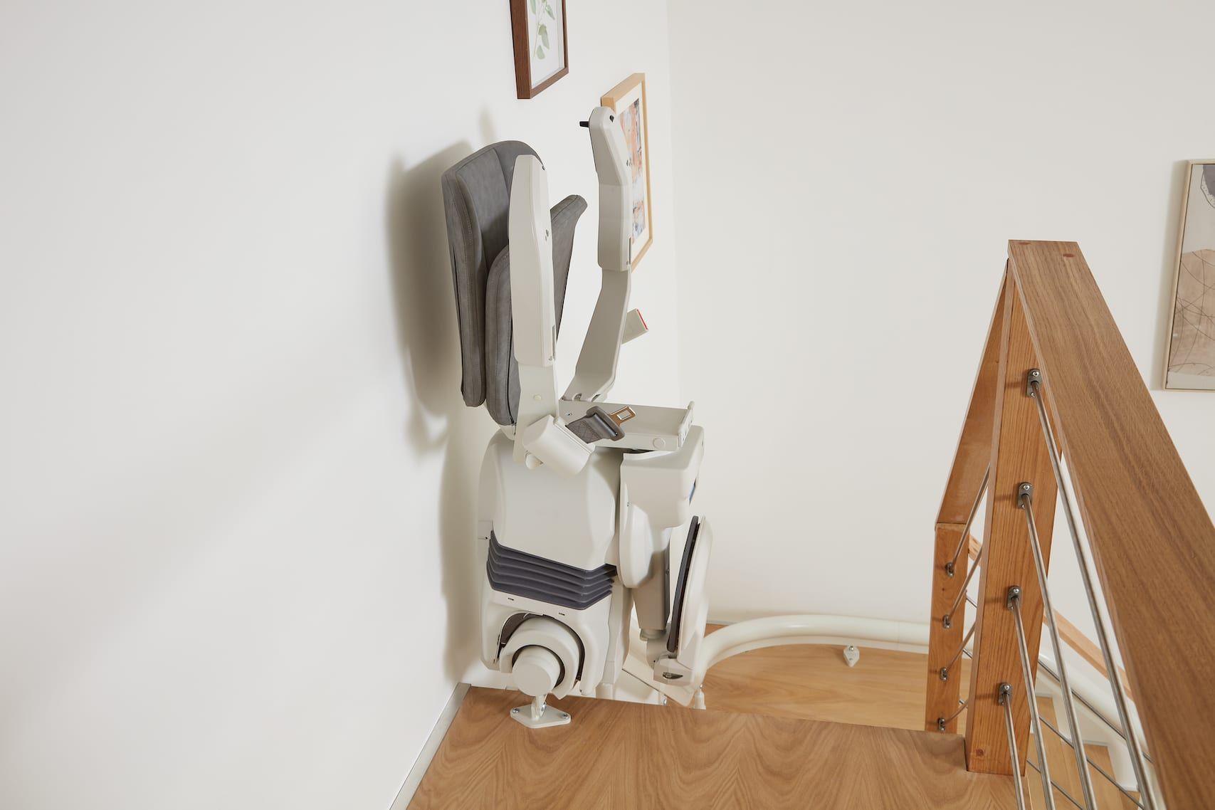 A stair lift, white and gray, on a wooden staircase in a house. A coat hangs on the lift.