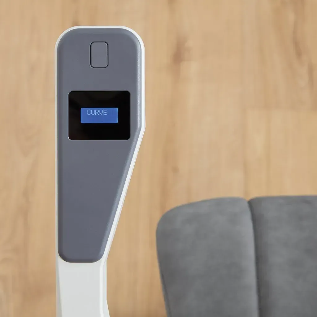 A modern, white and gray medical device with a small digital display stands against a light wood-paneled wall.