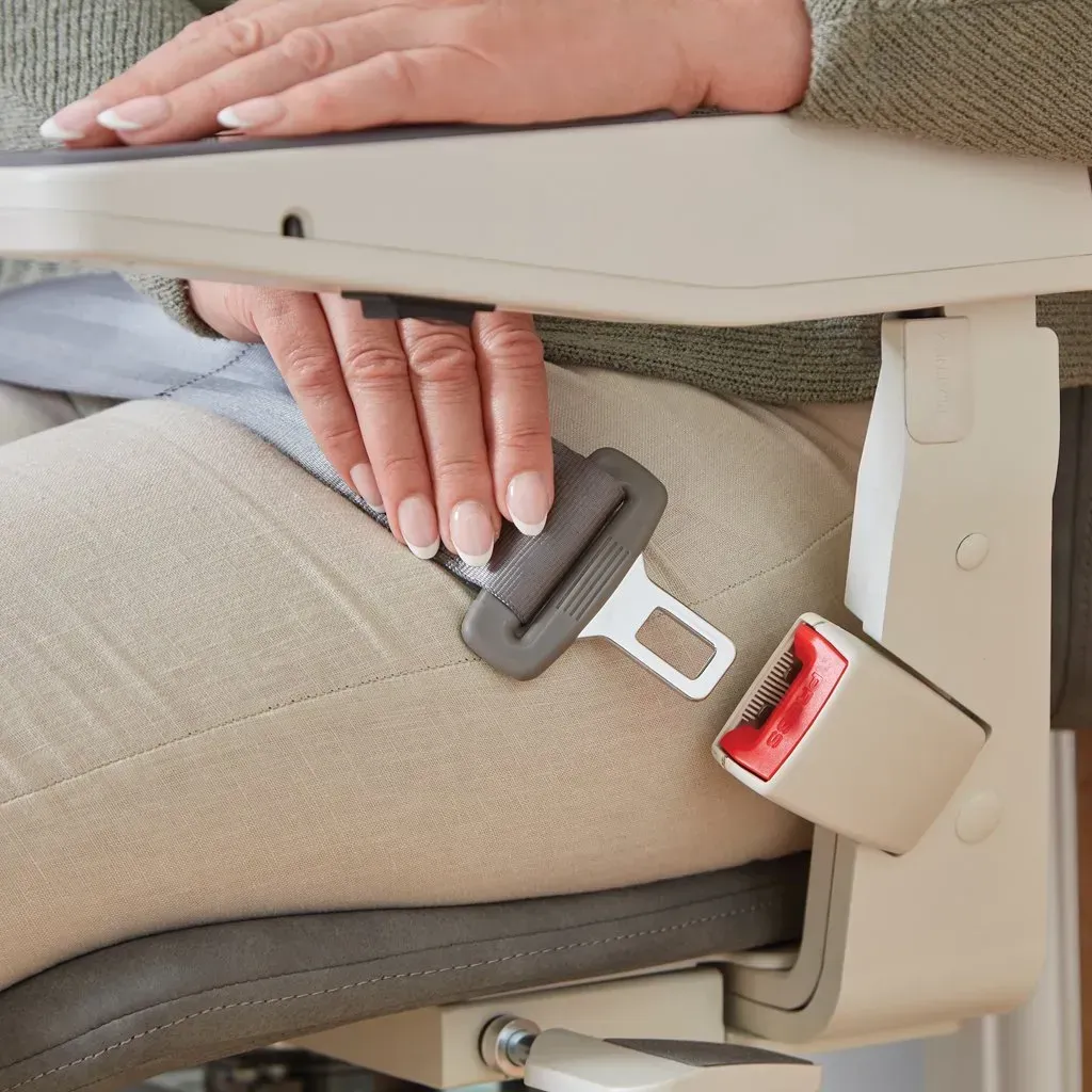 A person sitting in a stairlift, wearing a grey seatbelt secured by a red buckle.