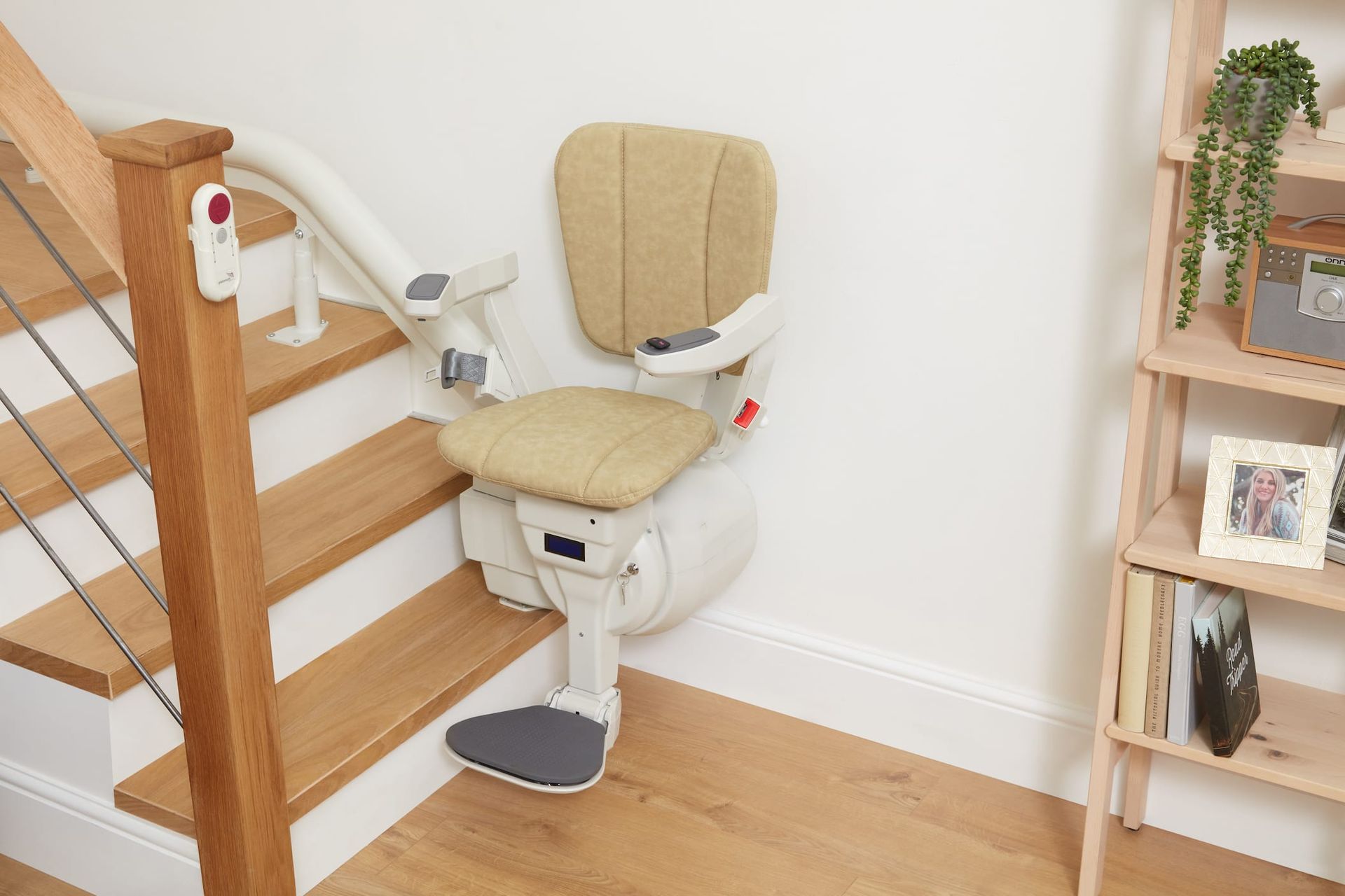 Stair lift chair mounted on a staircase. Beige chair, white railing, wooden stairs, and a shelf unit are visible.