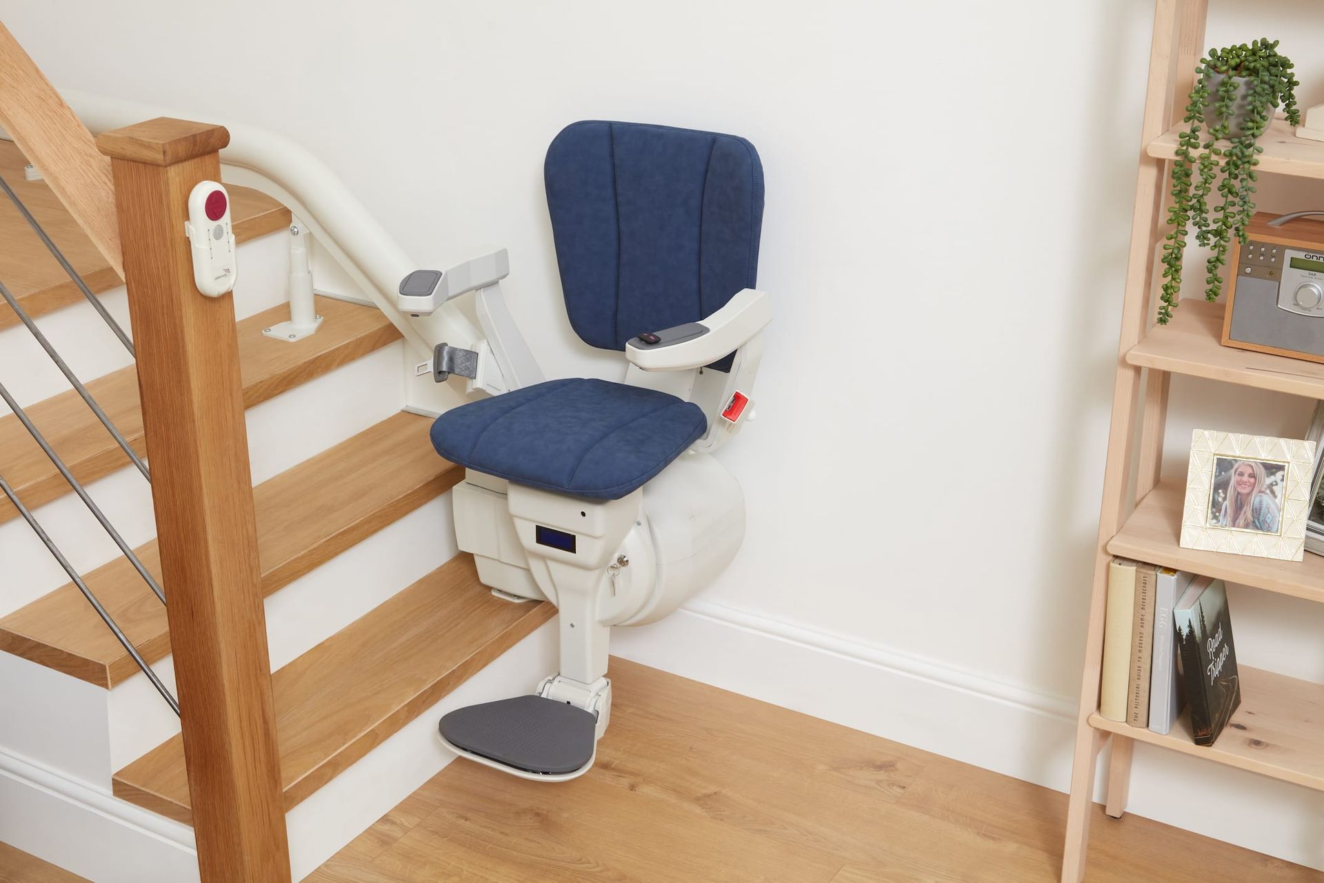 Stair lift with blue seat and back installed on wooden staircase, next to white wall and shelving unit.