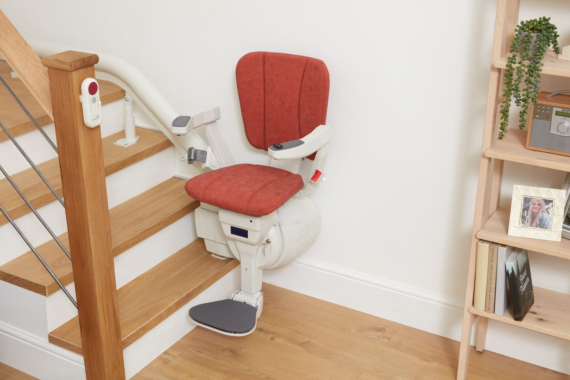 Stair lift installed on a wooden staircase with red seat and back against a white wall.