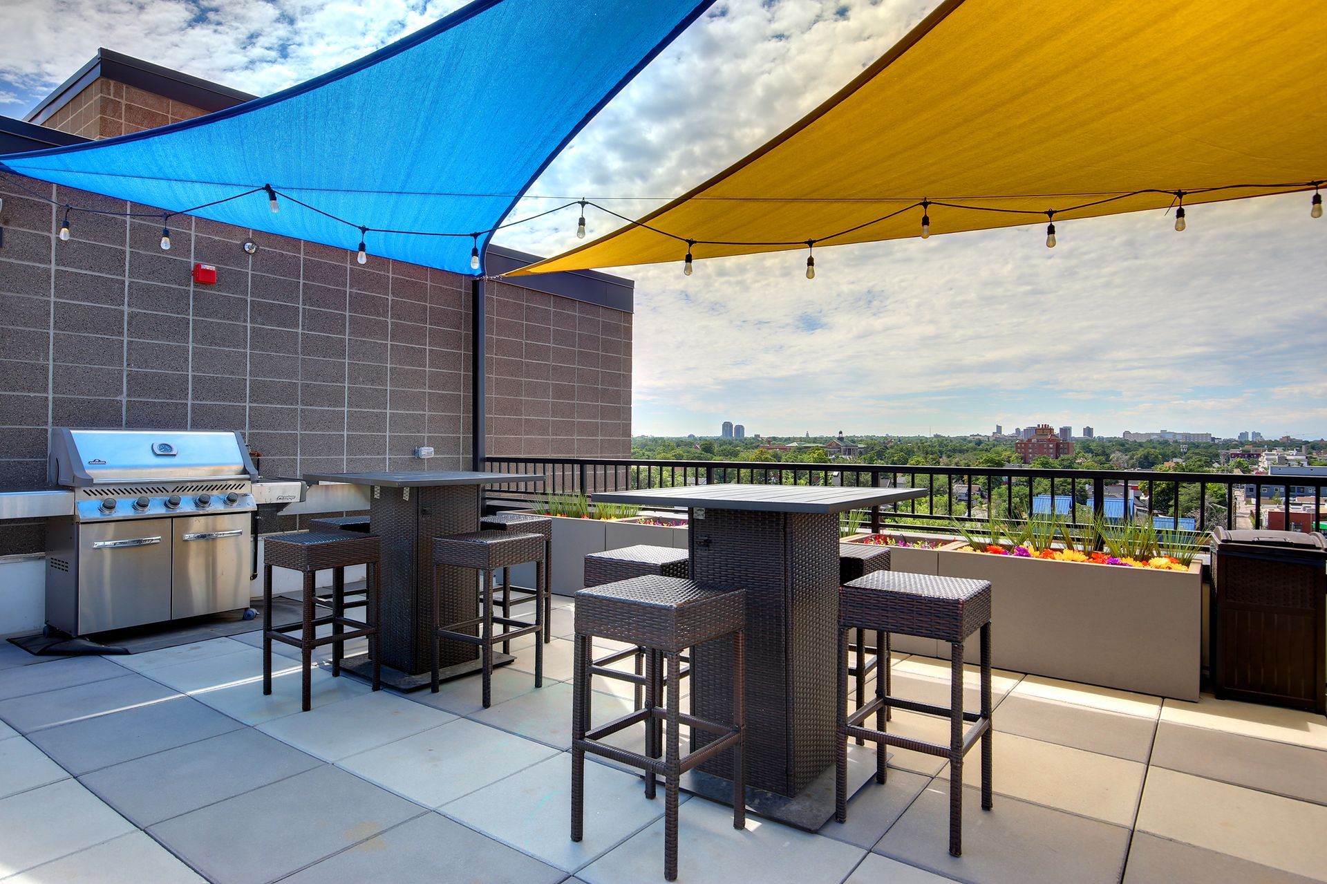 Rooftop patio with grill, high-top tables, stools, and shade sails in blue and yellow, overlooking a city skyline.